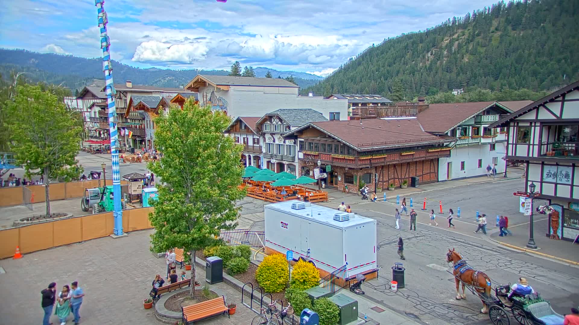 A Bavarian-style town square with people walking, a horse-drawn carriage, and outdoor seating under green umbrellas, all set against a backdrop of mountains under partly cloudy skies.