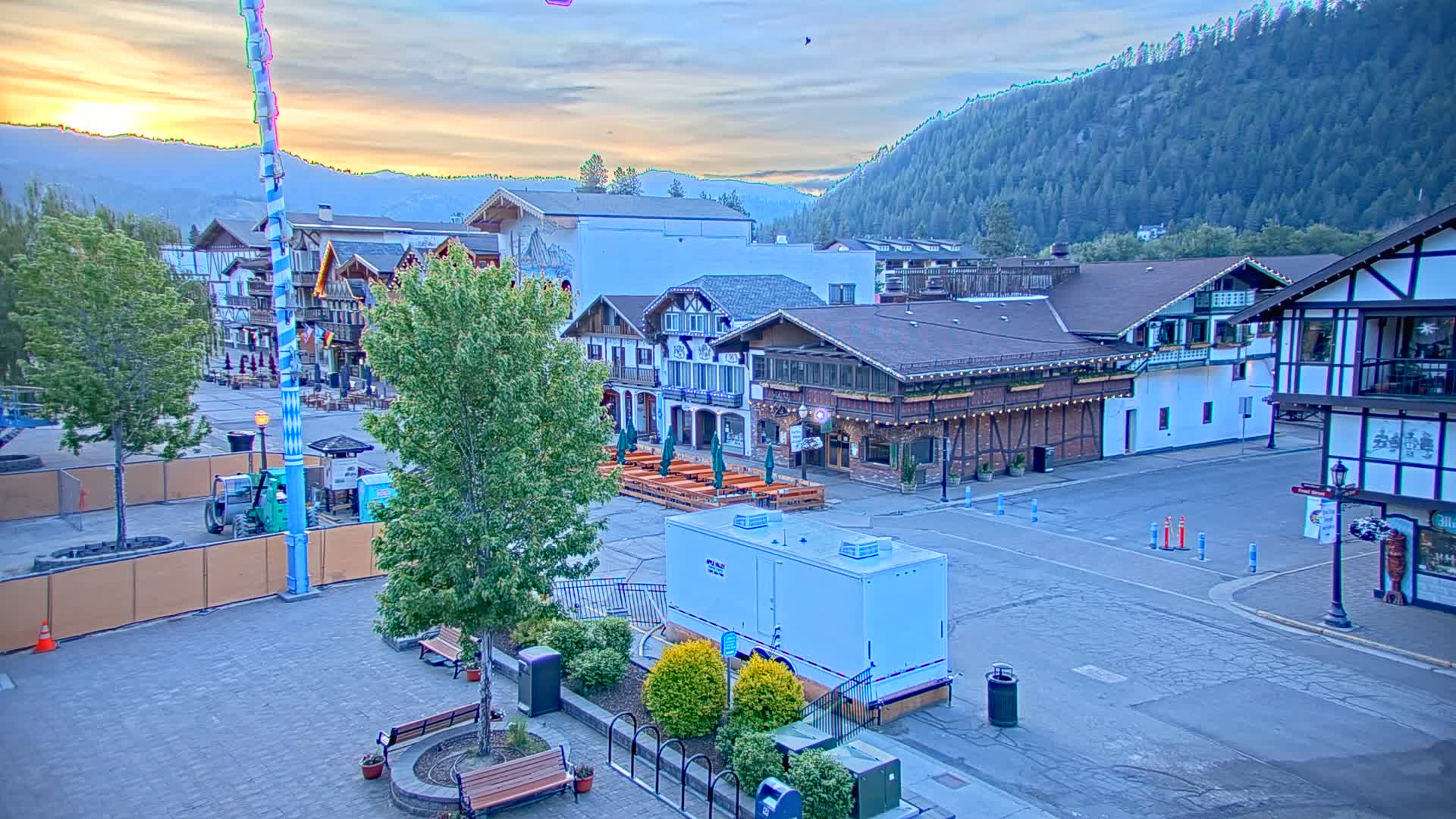 A town square with Bavarian-style buildings, outdoor seating areas, and a tall maypole, sits at the base of a mountain range under a sunrise sky.
