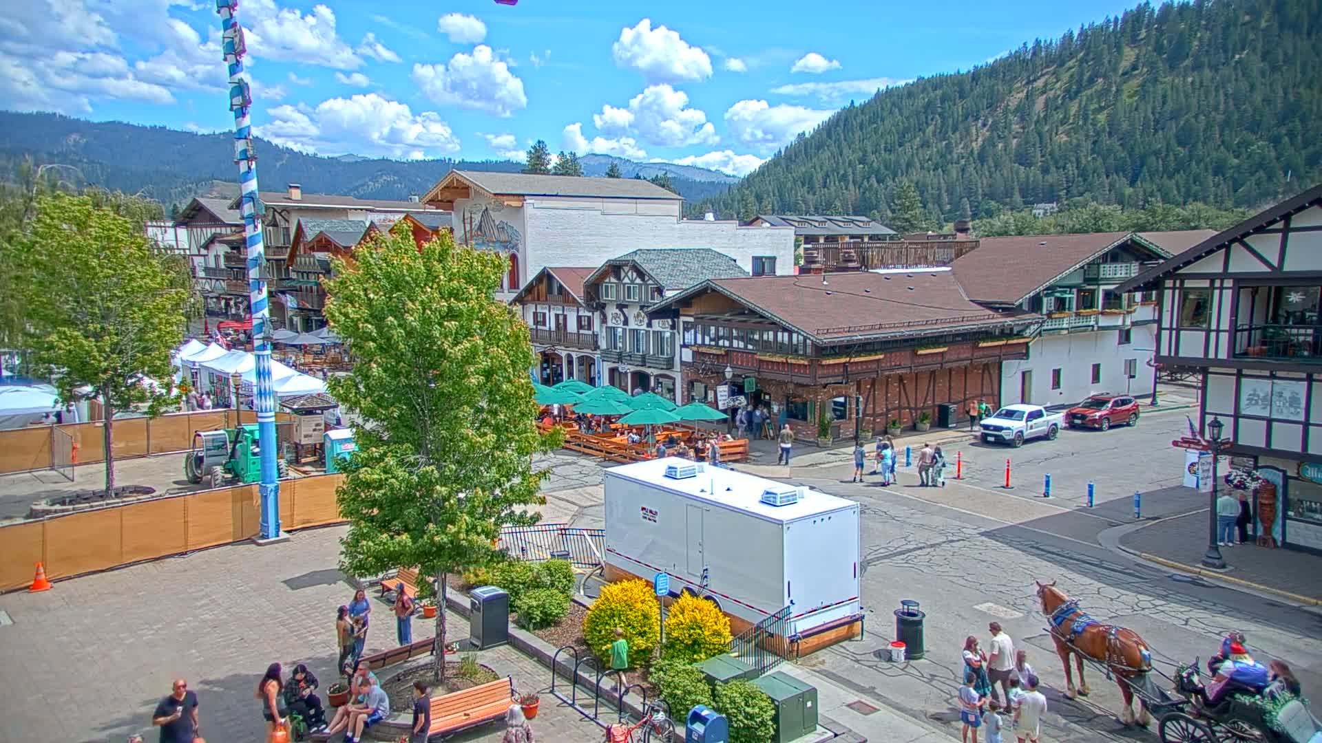 A Bavarian-style town square, under a partly sunny sky, features people gathered near outdoor seating, a horse-drawn carriage, and a tall maypole.