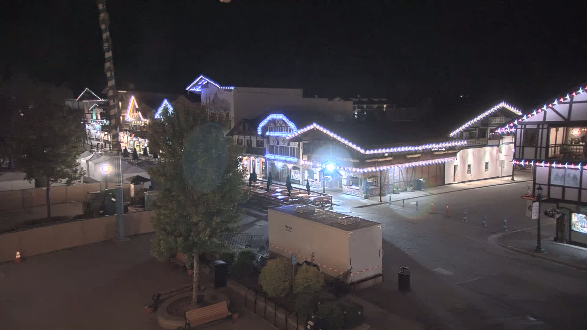 At night, a mostly empty town square with several buildings decorated with strings of lights is seen from an elevated perspective.
