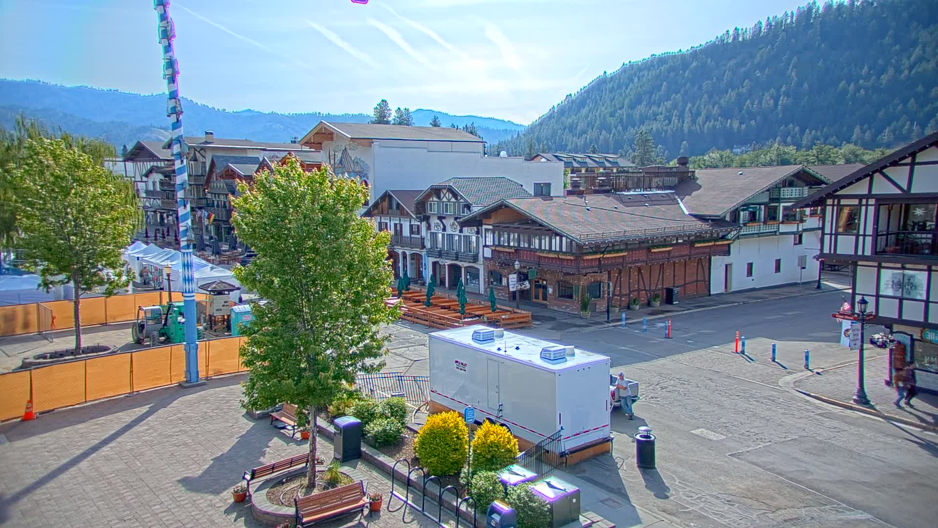 A Bavarian-style village square on a sunny day, surrounded by buildings, trees, and temporary event structures.