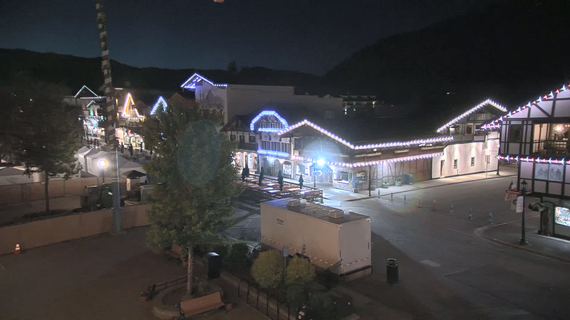 A nighttime, illuminated village square with several buildings decorated with string lights, a few trees, and a portable restroom.