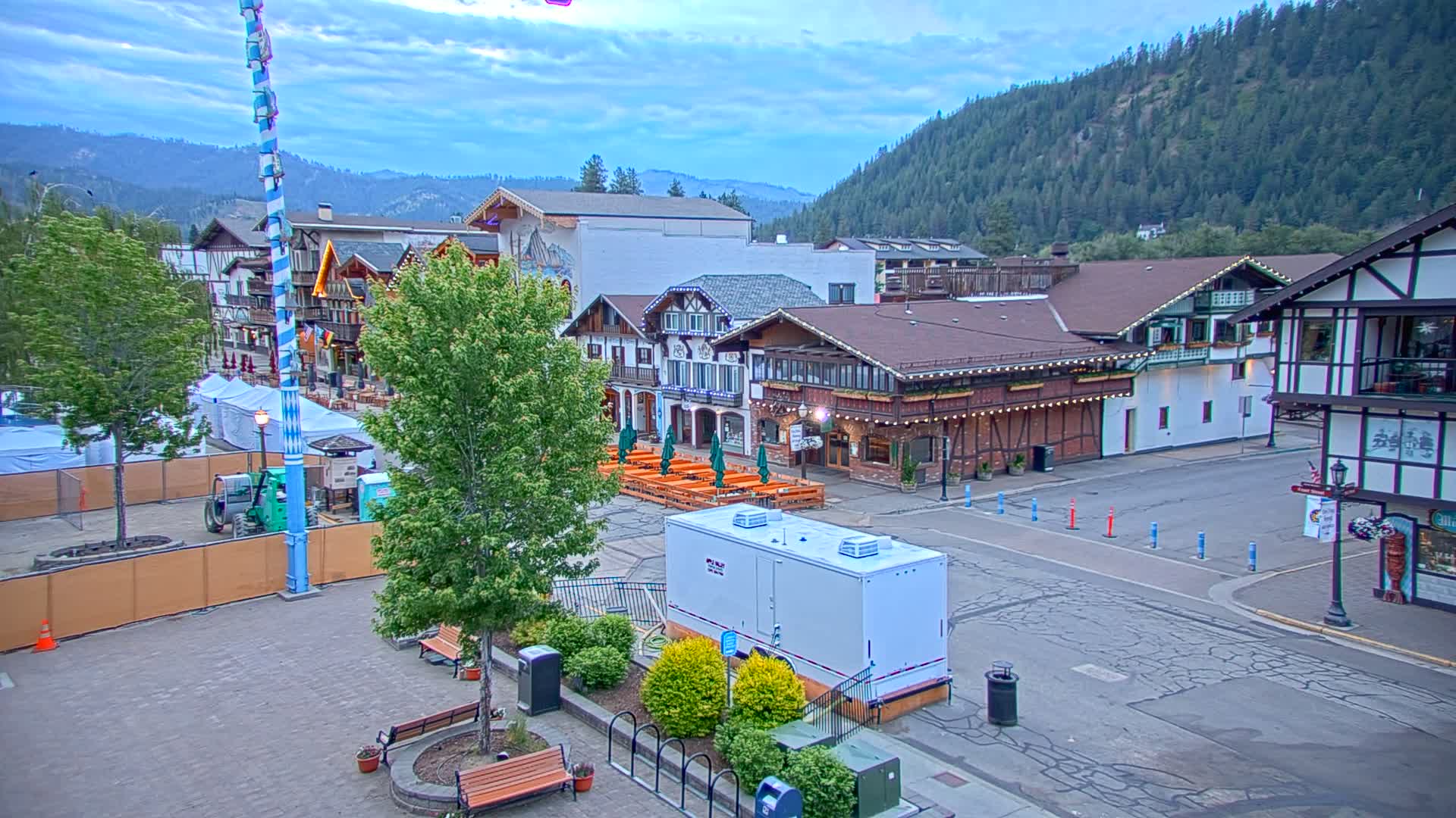A town square with Bavarian-style buildings under a partly cloudy sky, featuring outdoor seating areas and construction barriers.