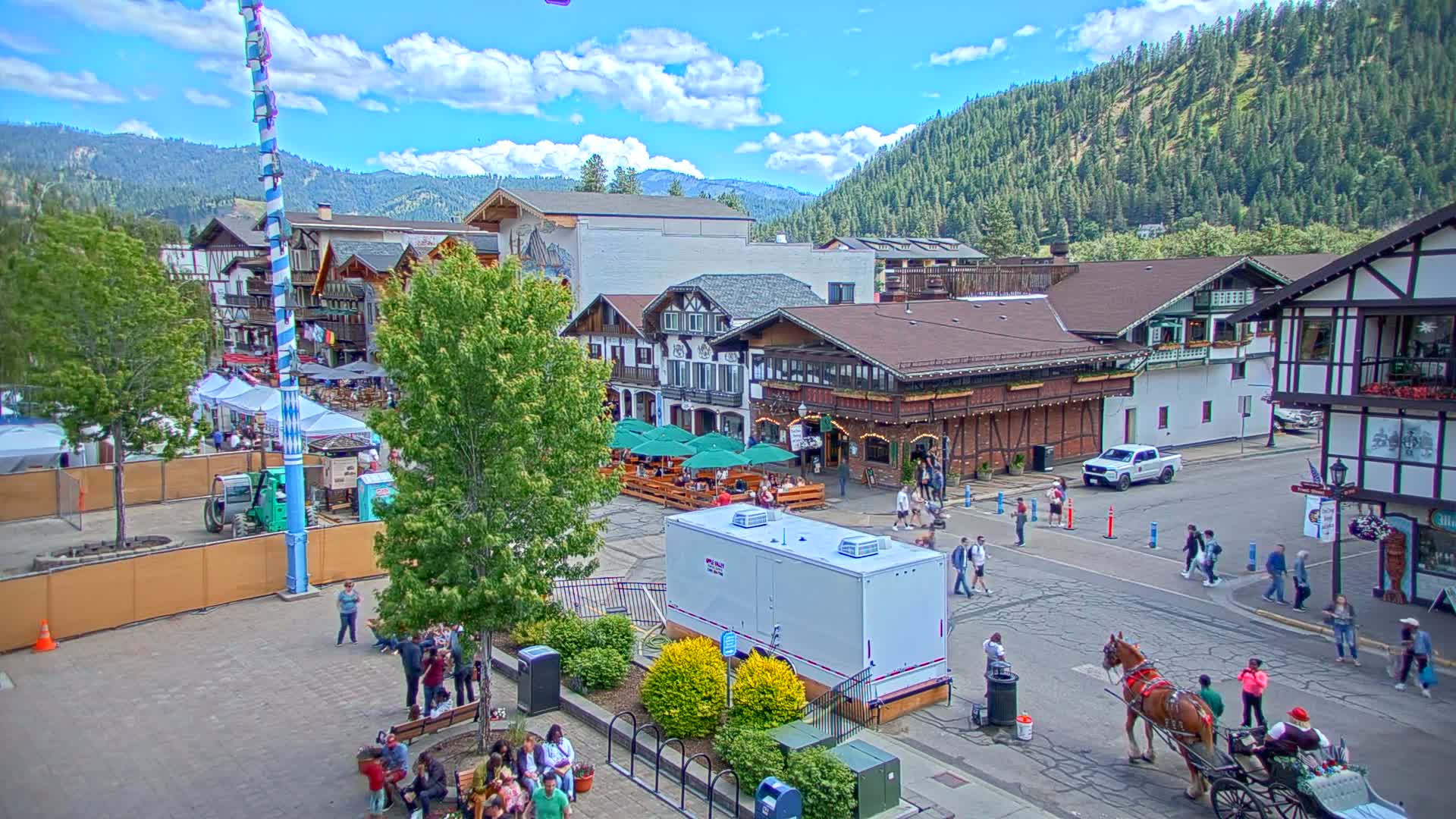 A Bavarian-style village square, under a partly cloudy sky, features people walking, outdoor seating areas at restaurants, a horse-drawn carriage, and a maypole.