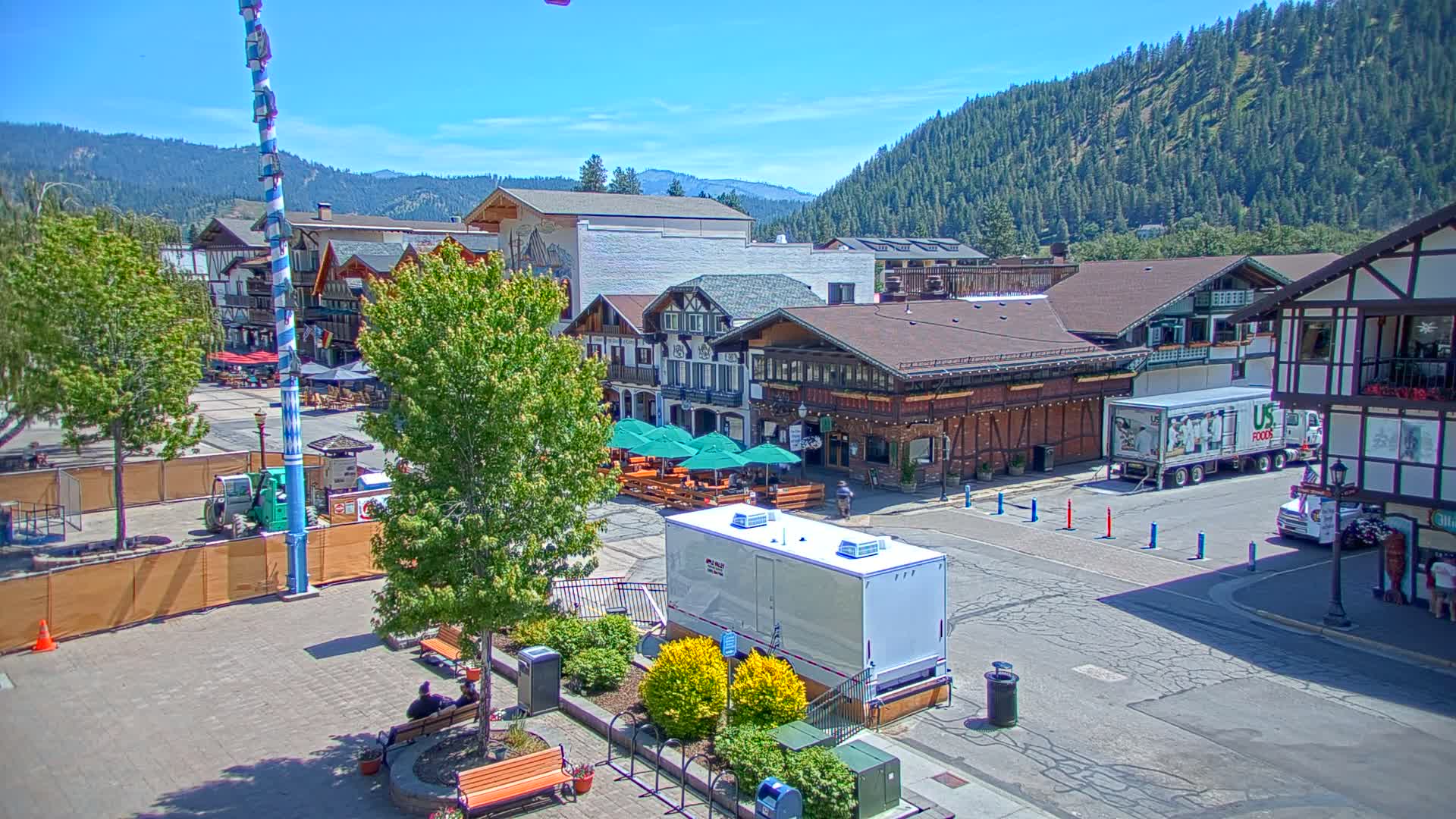 A town square with Bavarian-style buildings, outdoor seating areas, and a large maypole, all under a clear, sunny sky and surrounded by forested mountains.