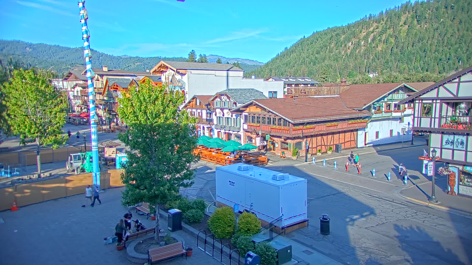 A sunny day reveals a town square with Bavarian-style buildings, outdoor seating, and a few pedestrians, all set against a backdrop of forested hills.