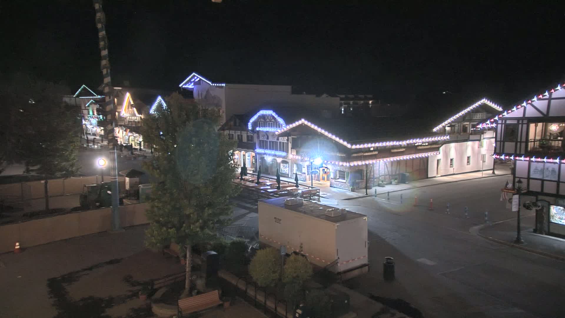A nighttime view of a street lined with festively lit buildings, a large trailer, and some trees, under a clear night sky.