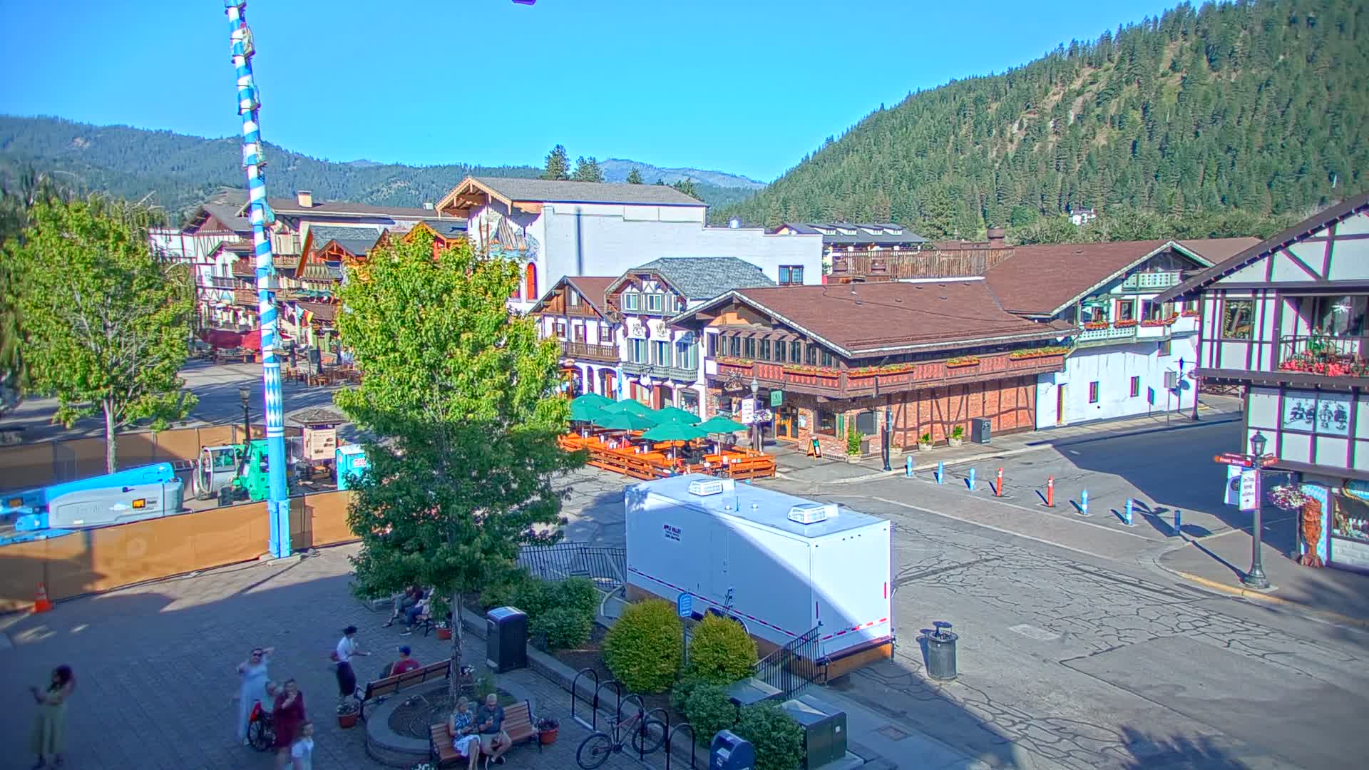 A sunny day reveals a Bavarian-style village square with several buildings, outdoor seating areas, and people strolling about, all nestled against a backdrop of forested hills.