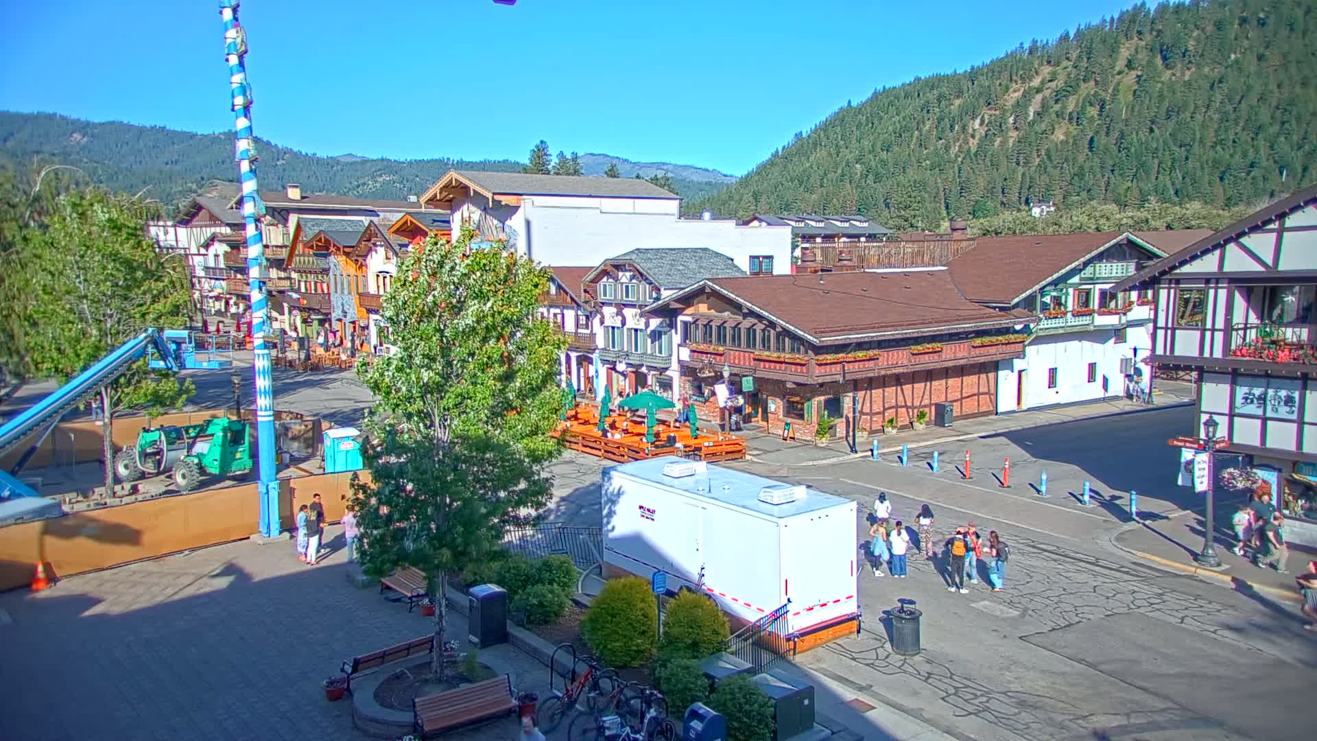 A Bavarian-style village square on a sunny day, with people walking, construction equipment, and outdoor seating areas in front of buildings against a backdrop of forested hills.