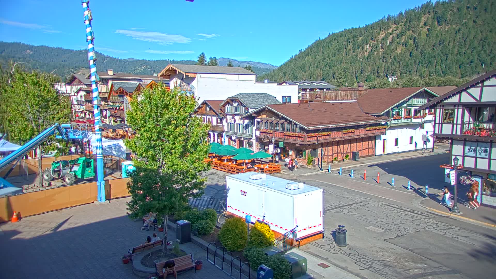 A sunny day reveals a Bavarian-style village square with several buildings, outdoor seating areas, construction equipment, and people walking around.