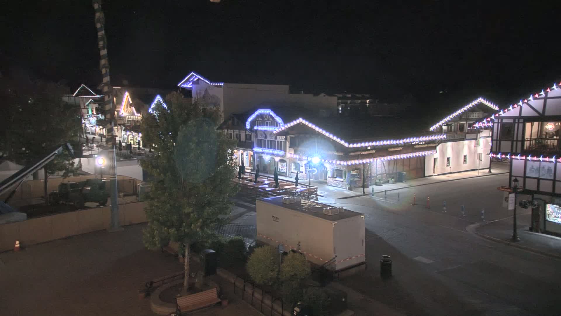 A nighttime view of a town square with several buildings decorated with string lights, a few trees, and a large portable restroom.