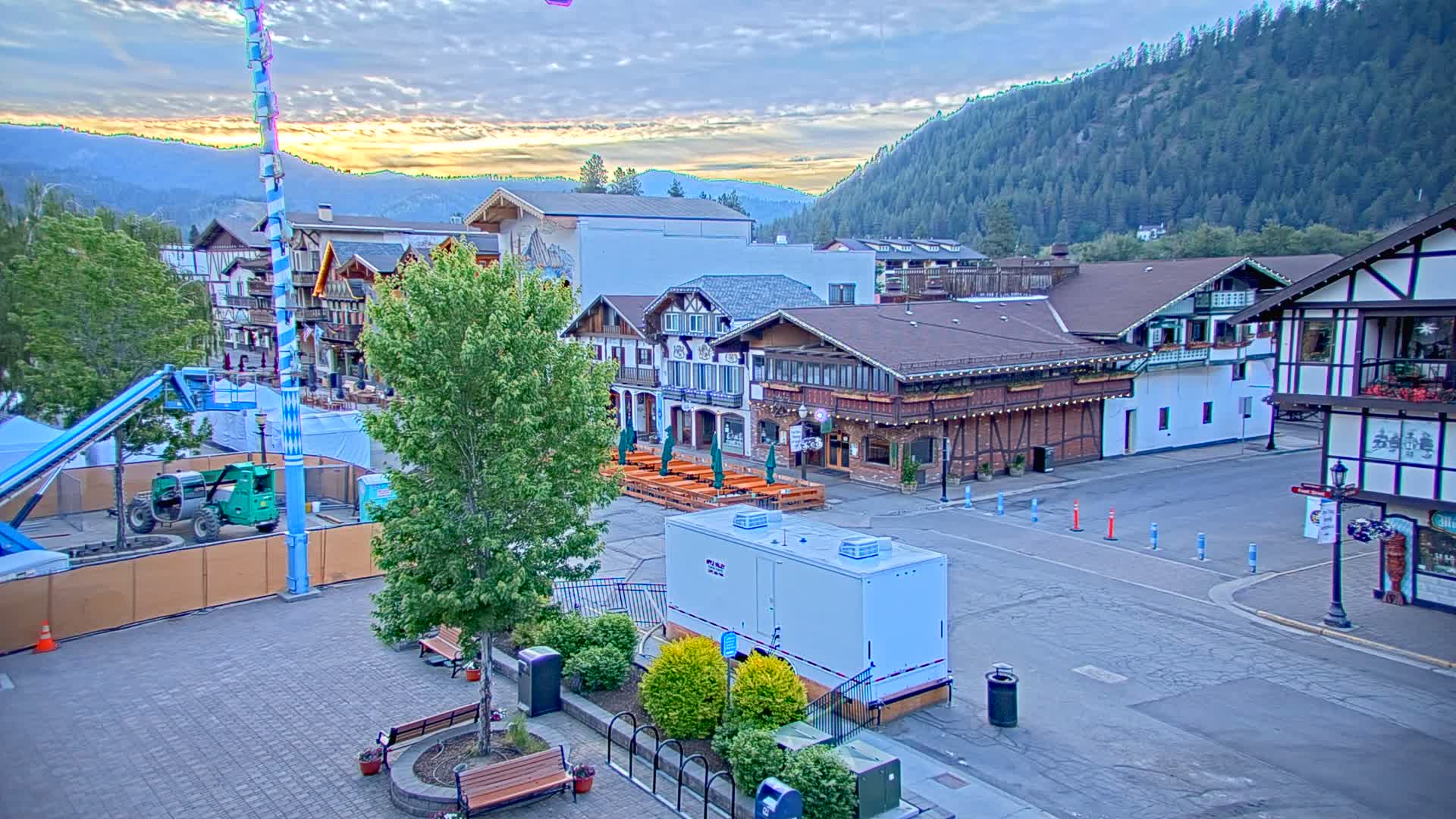 A town square with Bavarian-style buildings, outdoor seating areas, and construction equipment is visible under a partly cloudy sunrise sky, backed by a mountain range.