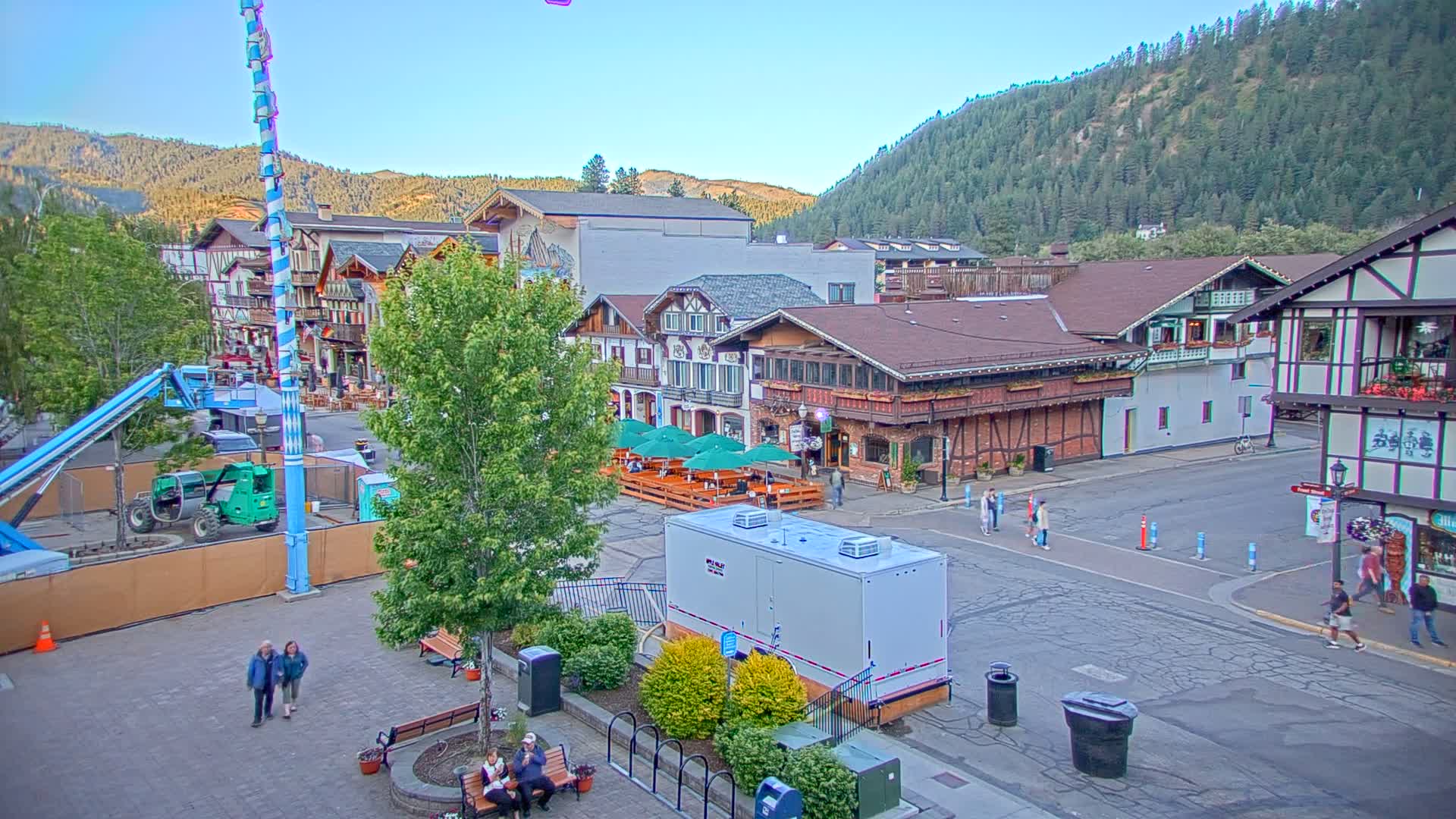 A town square with Bavarian-style buildings, a tall maypole, and several people walking around under a clear, sunny sky, backed by a mountain range.