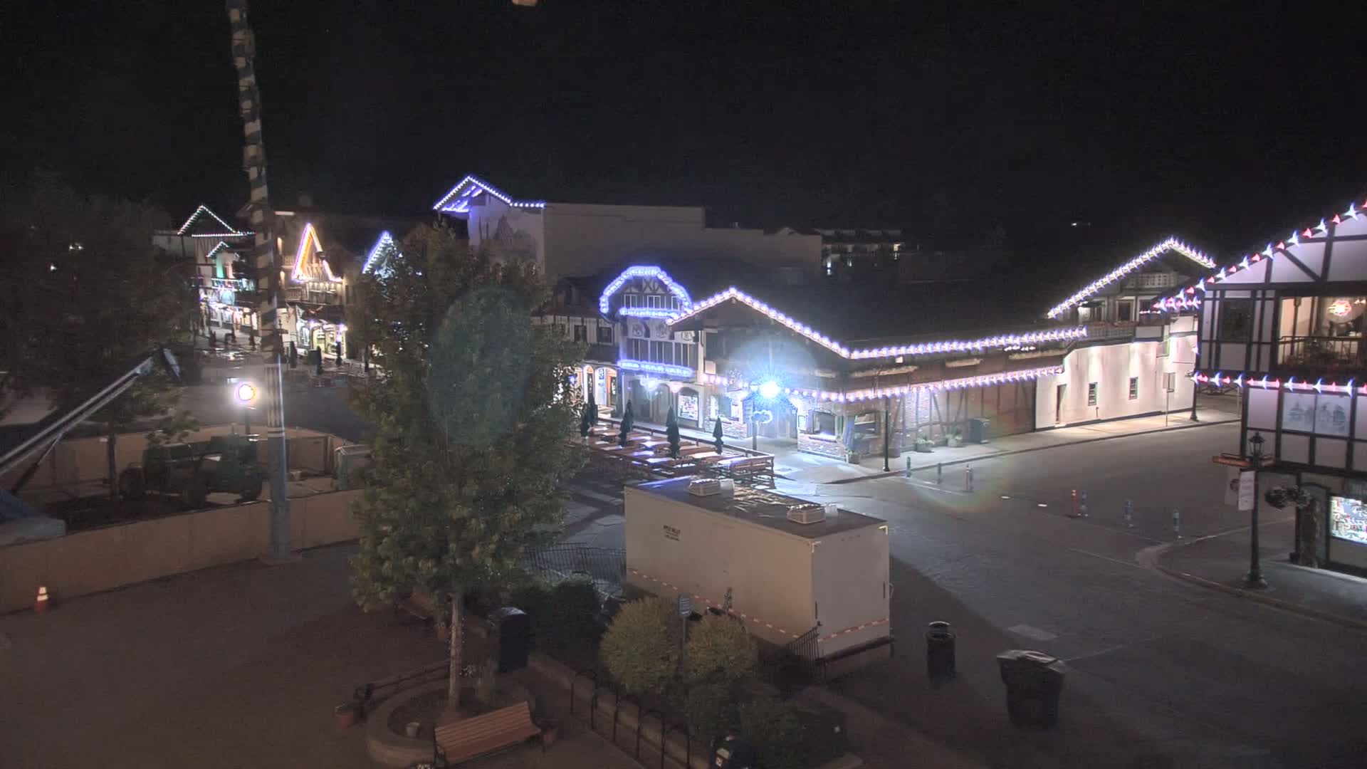 A nighttime, high-angle view shows a mostly empty street lined with decorated buildings, some with outdoor seating, under clear skies.