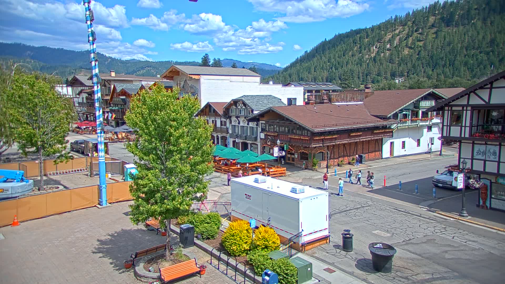 A Bavarian-style town square, under a partly sunny sky, features several buildings with outdoor seating, pedestrians, and a tall decorated pole.