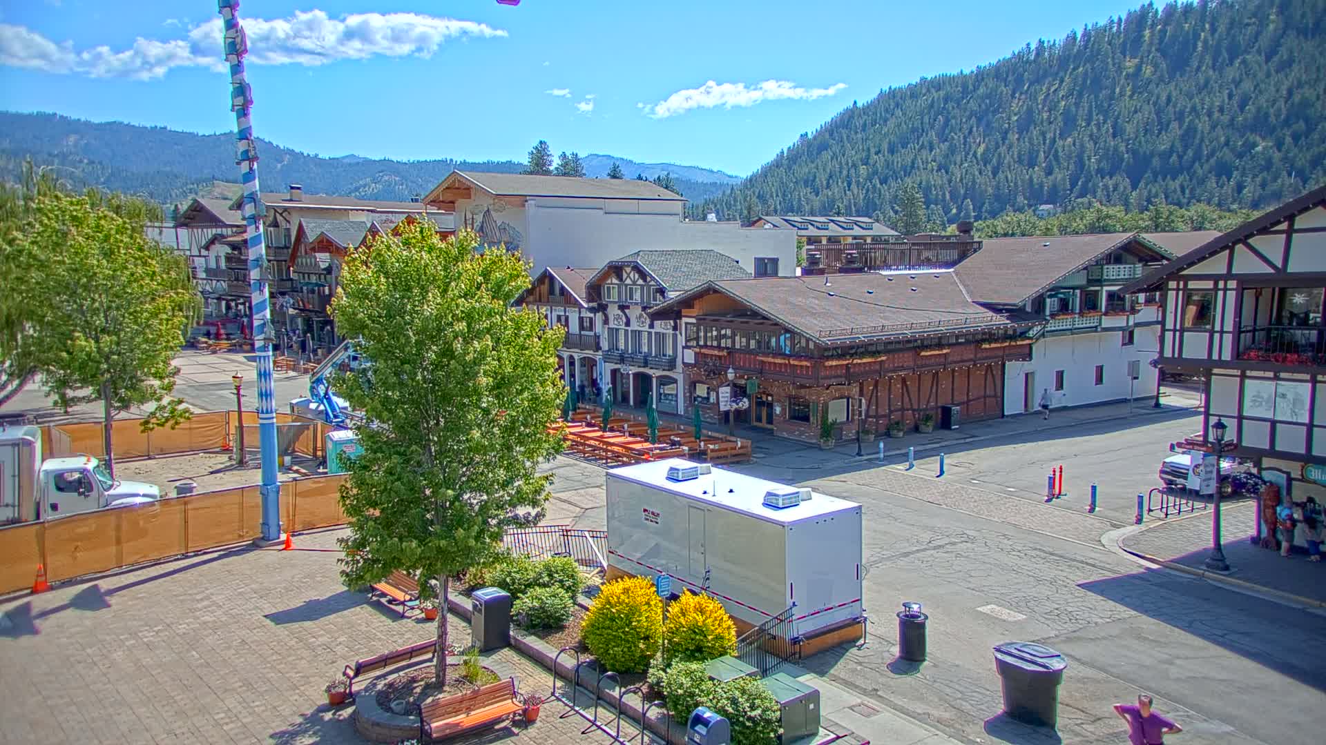 A sunny day reveals a town square with Bavarian-style buildings, trees, and outdoor seating, all set against a backdrop of a mountain range covered in lush green forest.