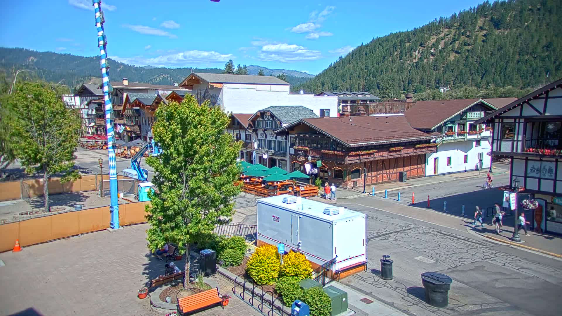 A sunny day in a Bavarian-style town square with a tall blue and white striped pole, several buildings with outdoor seating areas, and people walking on the street in front of a backdrop of a mountain range covered in green trees.