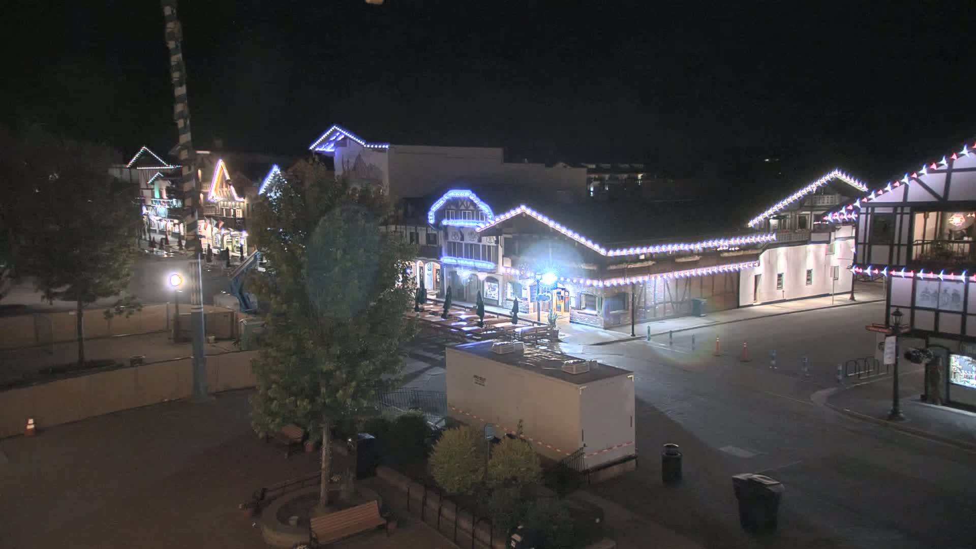 A nighttime view of a town square with several buildings decorated with string lights, trees, and some temporary construction barriers.