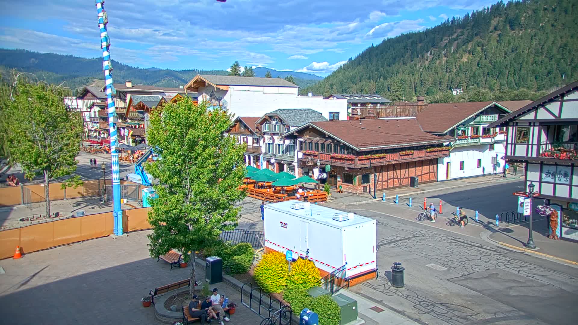 A town square with Bavarian-style buildings, outdoor seating areas, and a tall blue and white pole, under a partly sunny sky with mountains in the background.