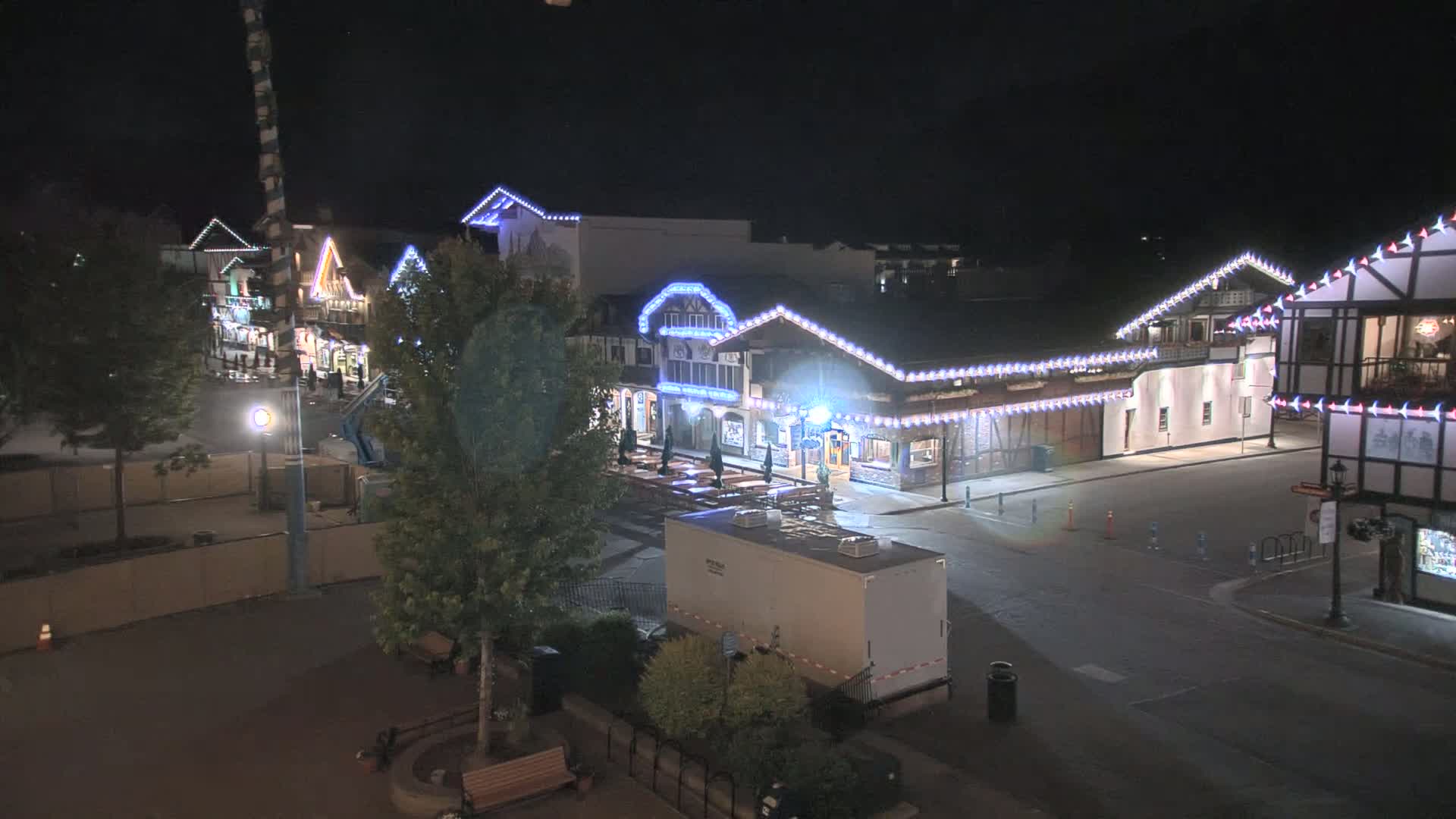 A nighttime, outdoor view of a row of buildings decorated with string lights, a mostly empty street, and a portable restroom.