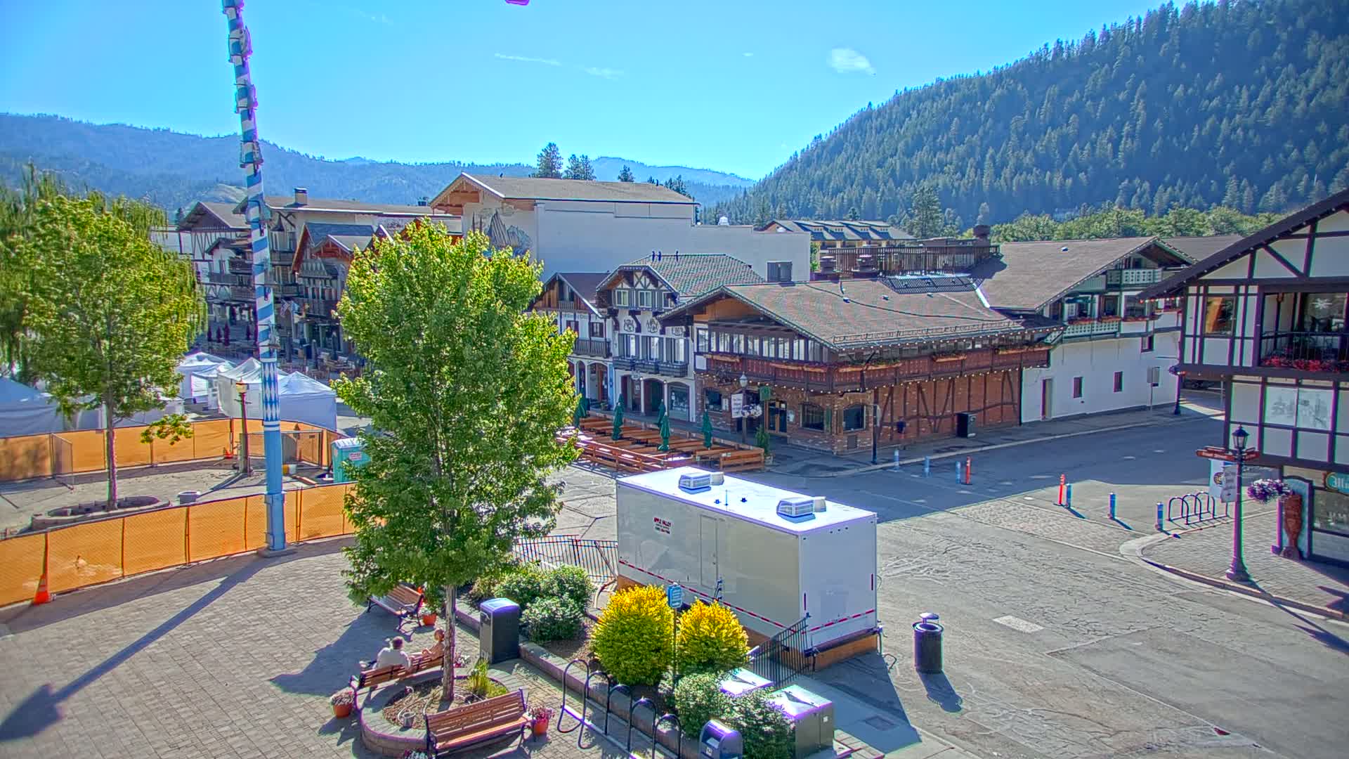 A town square with Bavarian-style buildings, a tall maypole, and outdoor seating is under a clear, sunny sky, with a forested mountain range in the background.