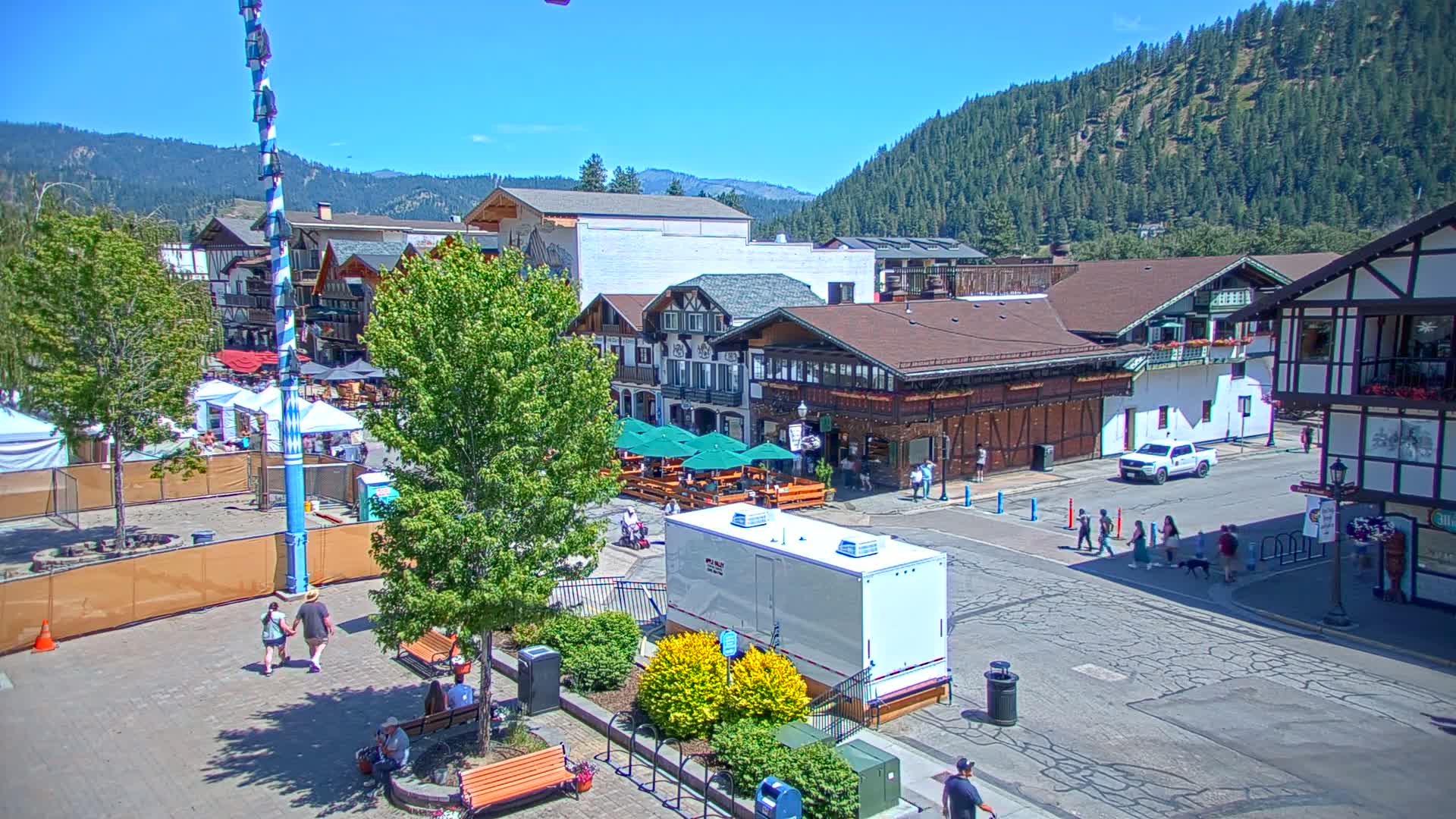 A sunny day in a Bavarian-style village town square with people walking around, several buildings, outdoor seating areas, and a tall decorated pole.