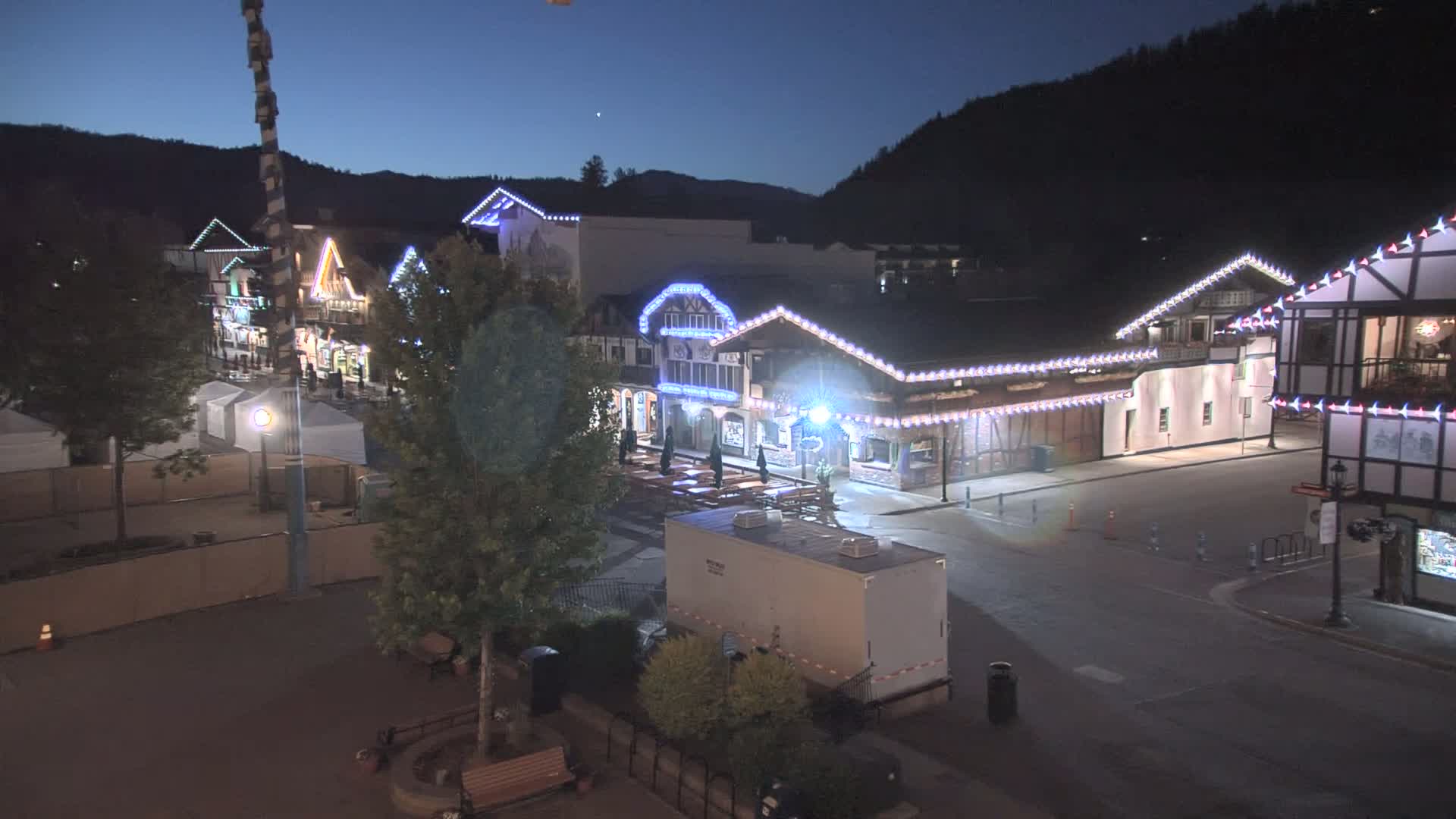 A nighttime, outdoor view of a town square with several buildings decorated with string lights, trees, and a portable restroom.