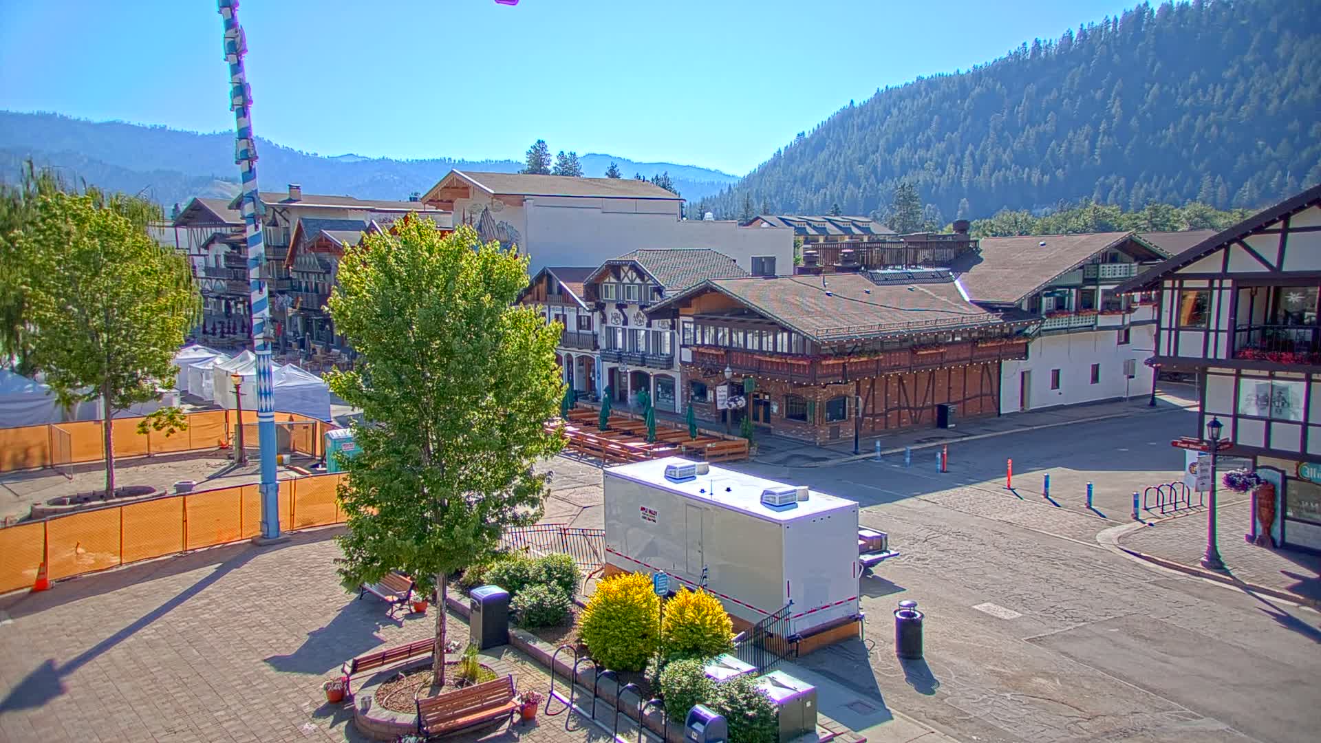 A town square with several buildings in a Bavarian style, a tall maypole, and a portable restroom, all under a clear, sunny sky, with a mountain range in the background.