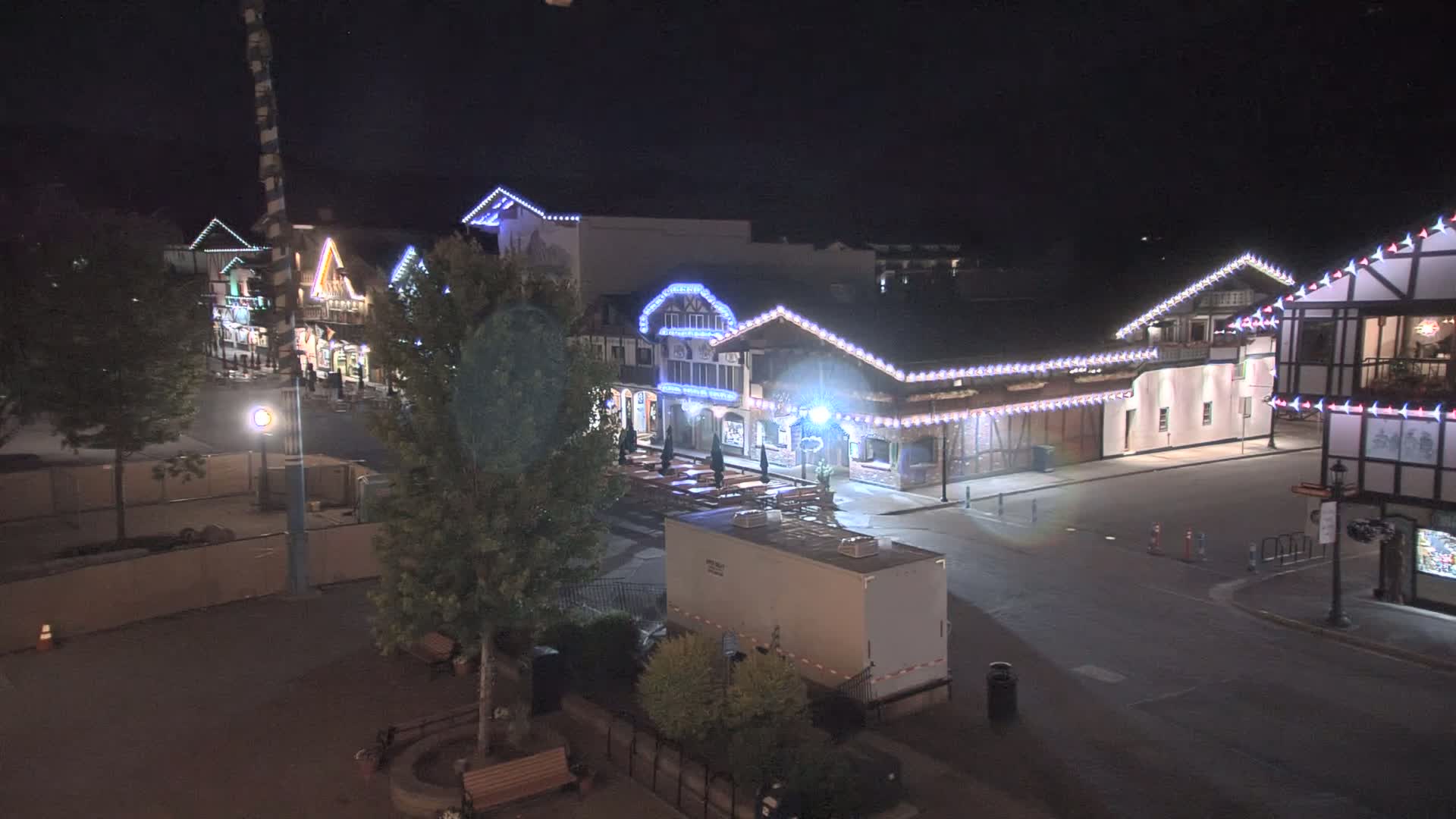 A nighttime, high-angle view of a street lined with illuminated buildings of a Bavarian style, featuring outdoor seating and a portable restroom.