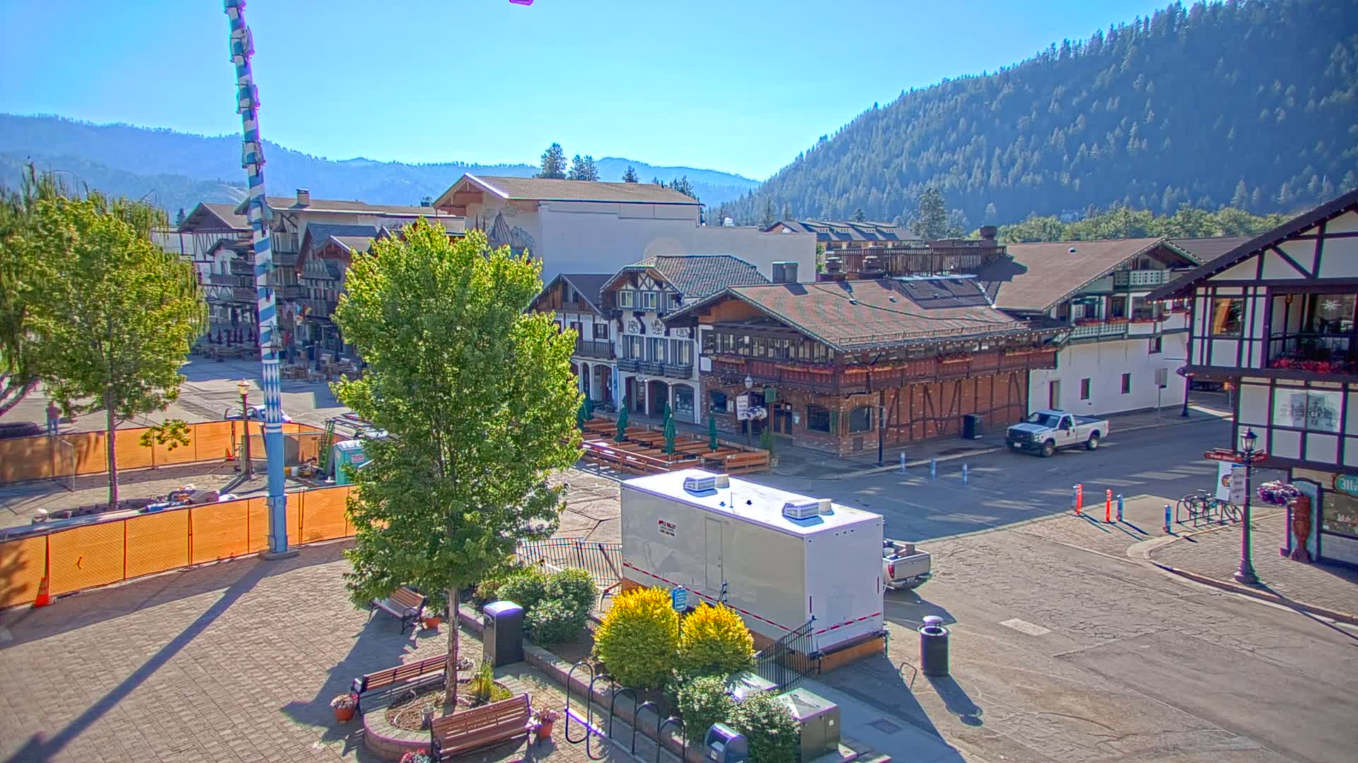 A town square with Bavarian-style buildings, green trees, and a clear sunny day, situated in a valley with forested hills in the background.