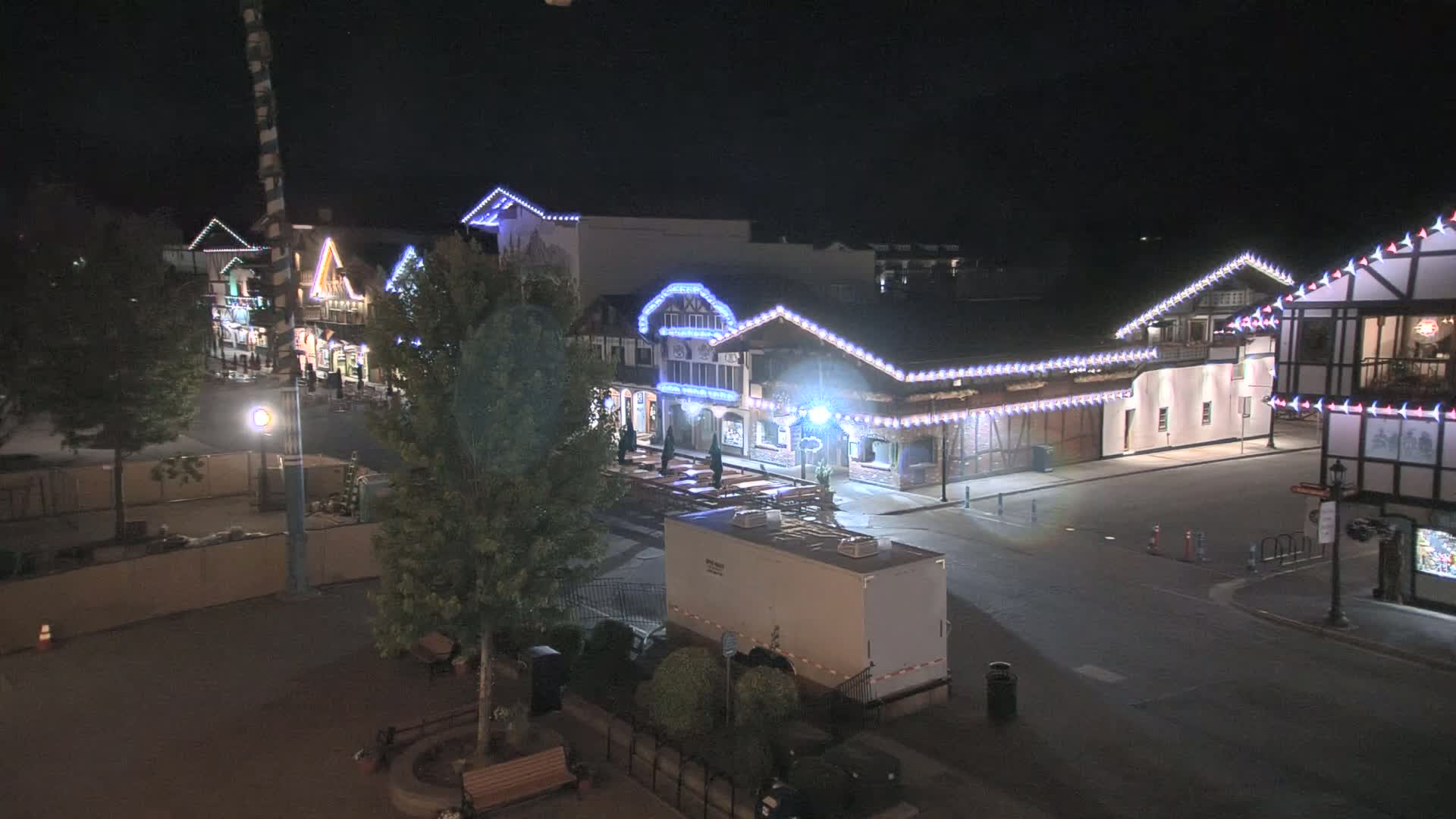 A nighttime view of a village-like street with several buildings decorated with string lights, mostly empty, under a clear, dark sky.