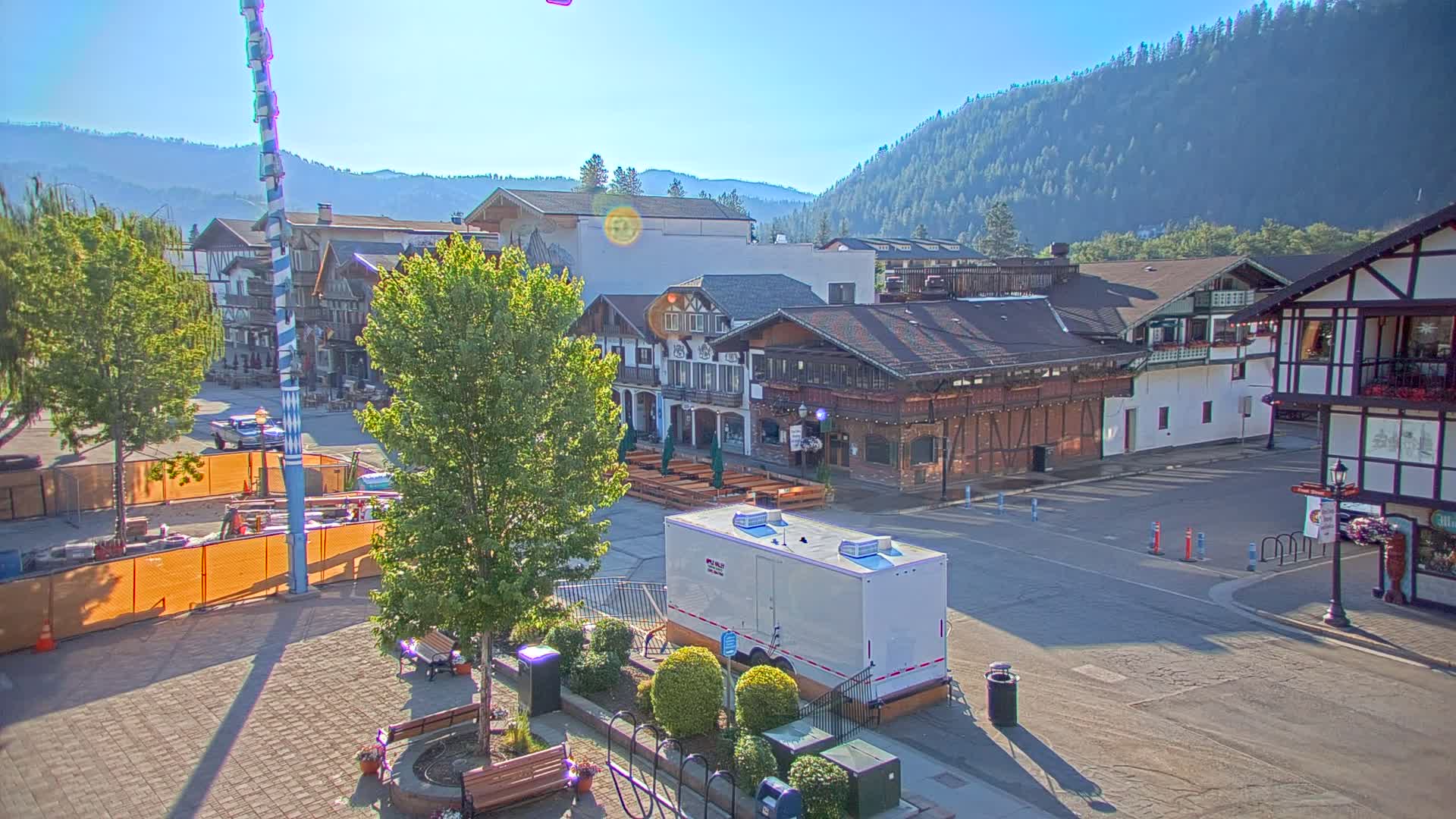 A sunny day reveals a town square with Bavarian-style buildings, a tall maypole, and a portable restroom, all set against a backdrop of forested hills.