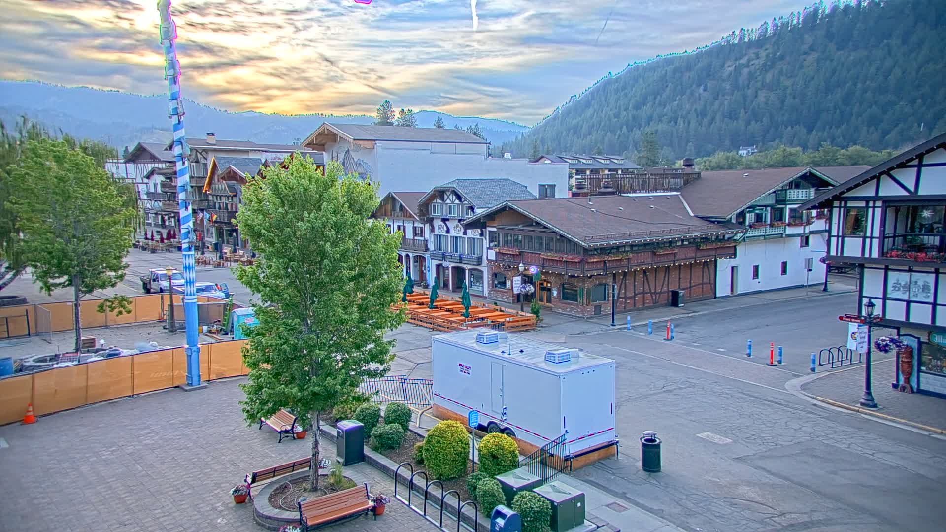 A mostly empty town square with Bavarian-style buildings, a tall maypole, and a portable restroom, all under a partly cloudy sky with mountains in the background.