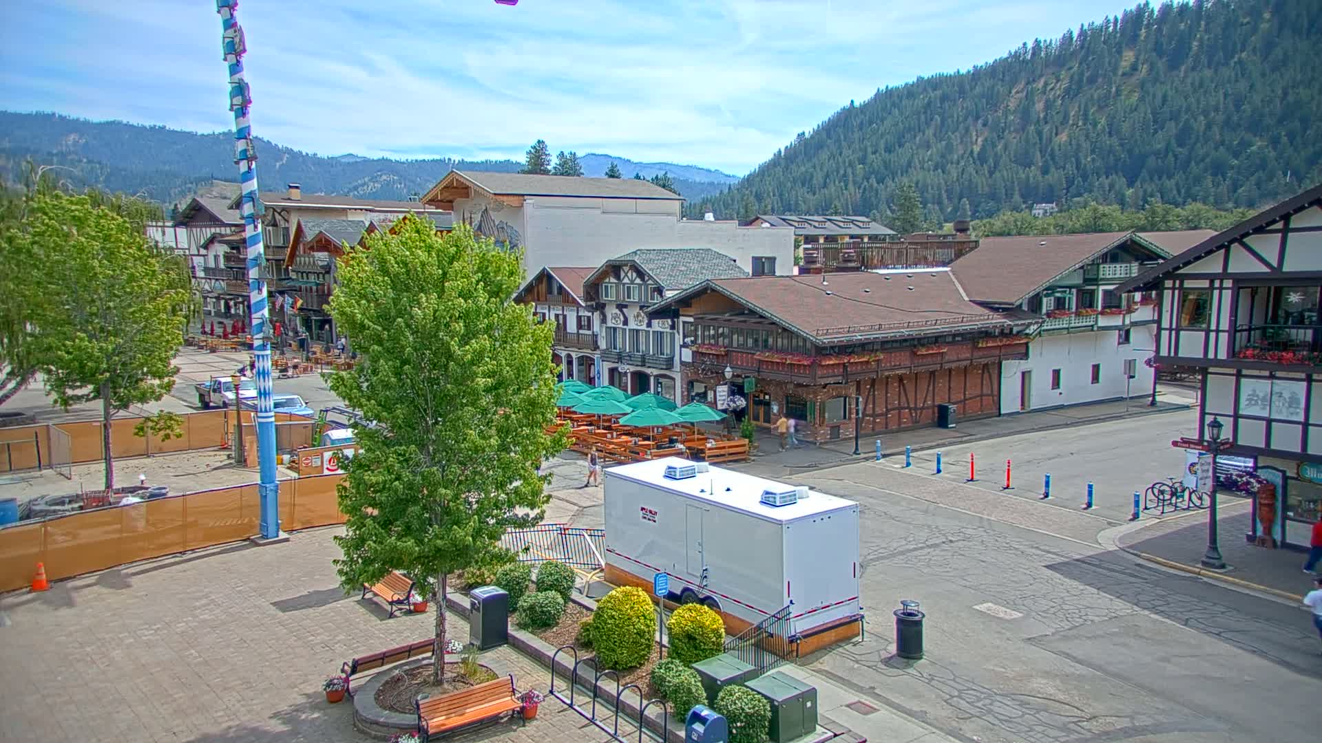 A town square with Bavarian-style buildings, outdoor seating, and a tall maypole under a sunny sky, surrounded by forested hills.