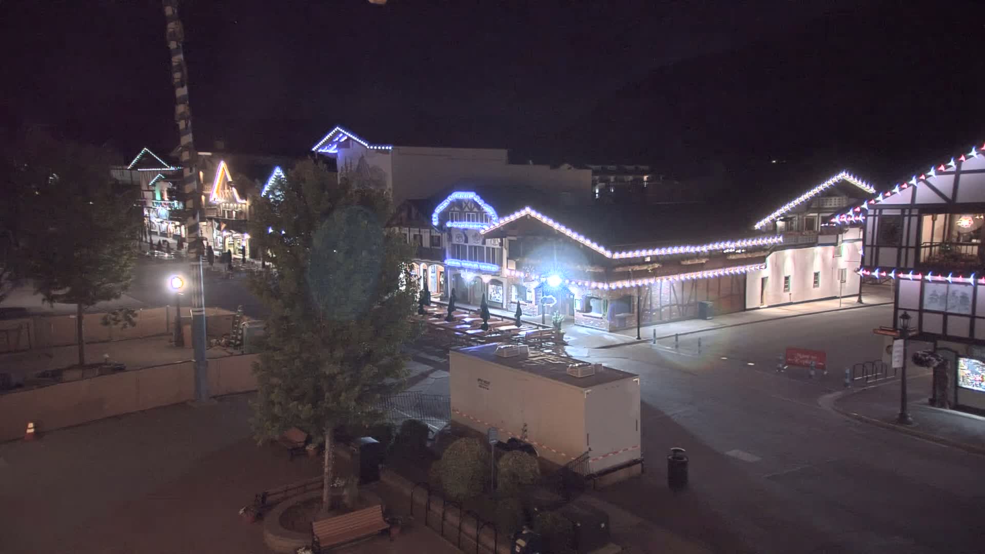 A nighttime, outdoor view of a mostly empty town square with several buildings decorated with string lights, a large Maypole, and a few trees.
