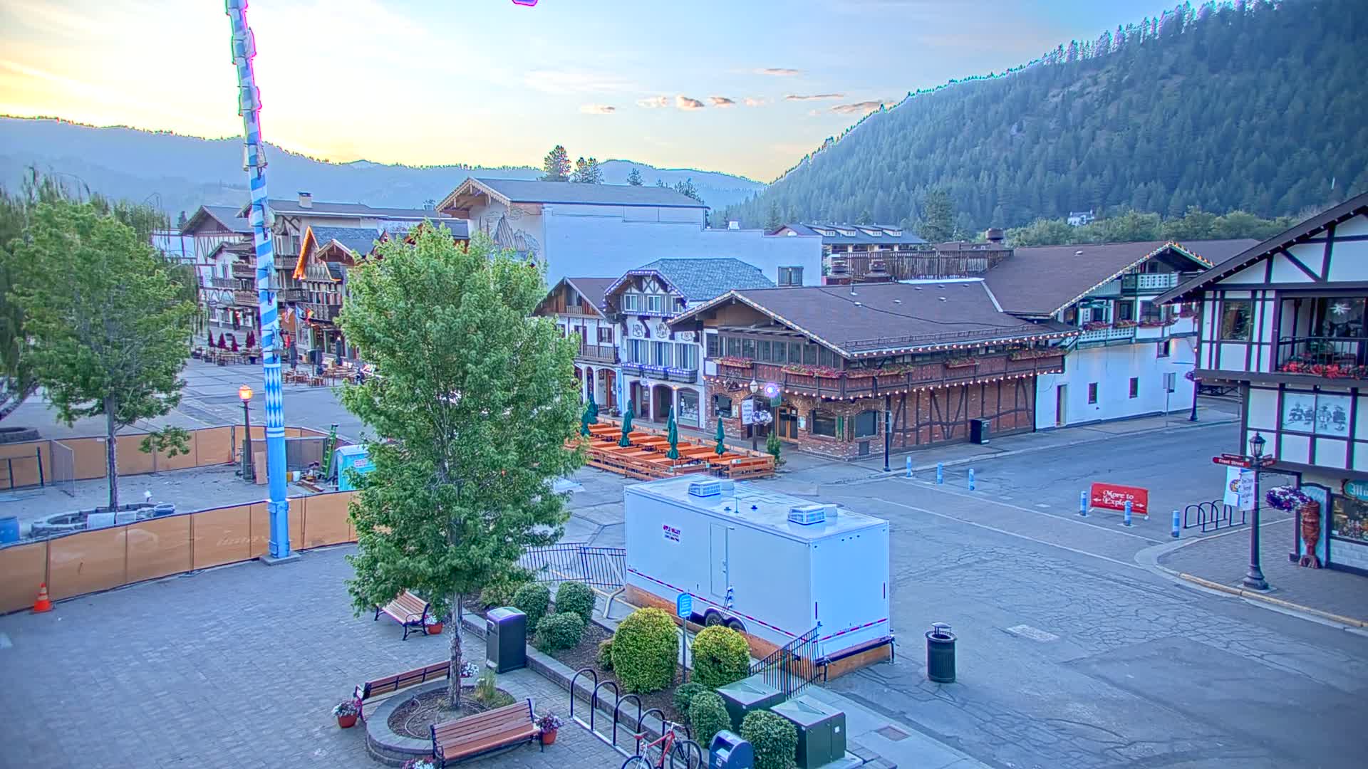 A town square with Bavarian-style buildings, outdoor seating areas, and lush green trees, under a clear twilight sky with mountains in the background.