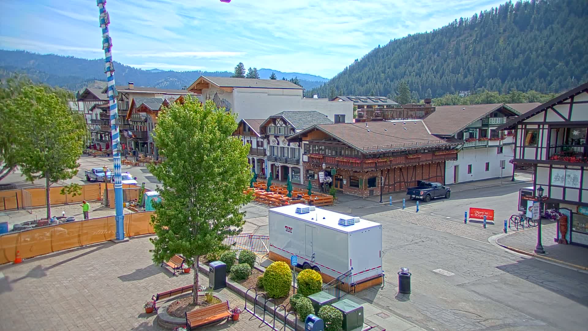 A Bavarian-style town square, under sunny skies, features buildings with outdoor seating, a tall maypole, and a portable restroom.