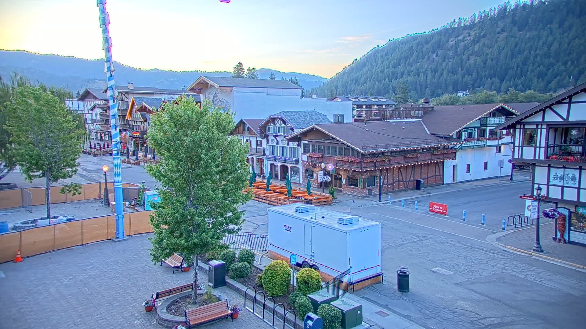 A paved town square with Bavarian-style buildings, outdoor seating, and a tall, striped pole under a clear, dawn sky against a backdrop of forested hills.