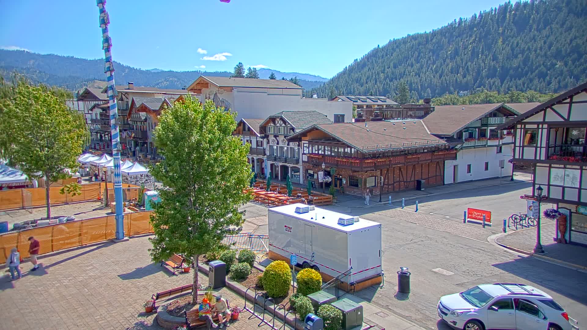 A town square with Bavarian-style buildings, a tall maypole, and a few people milling around on a sunny day, with mountains in the background.