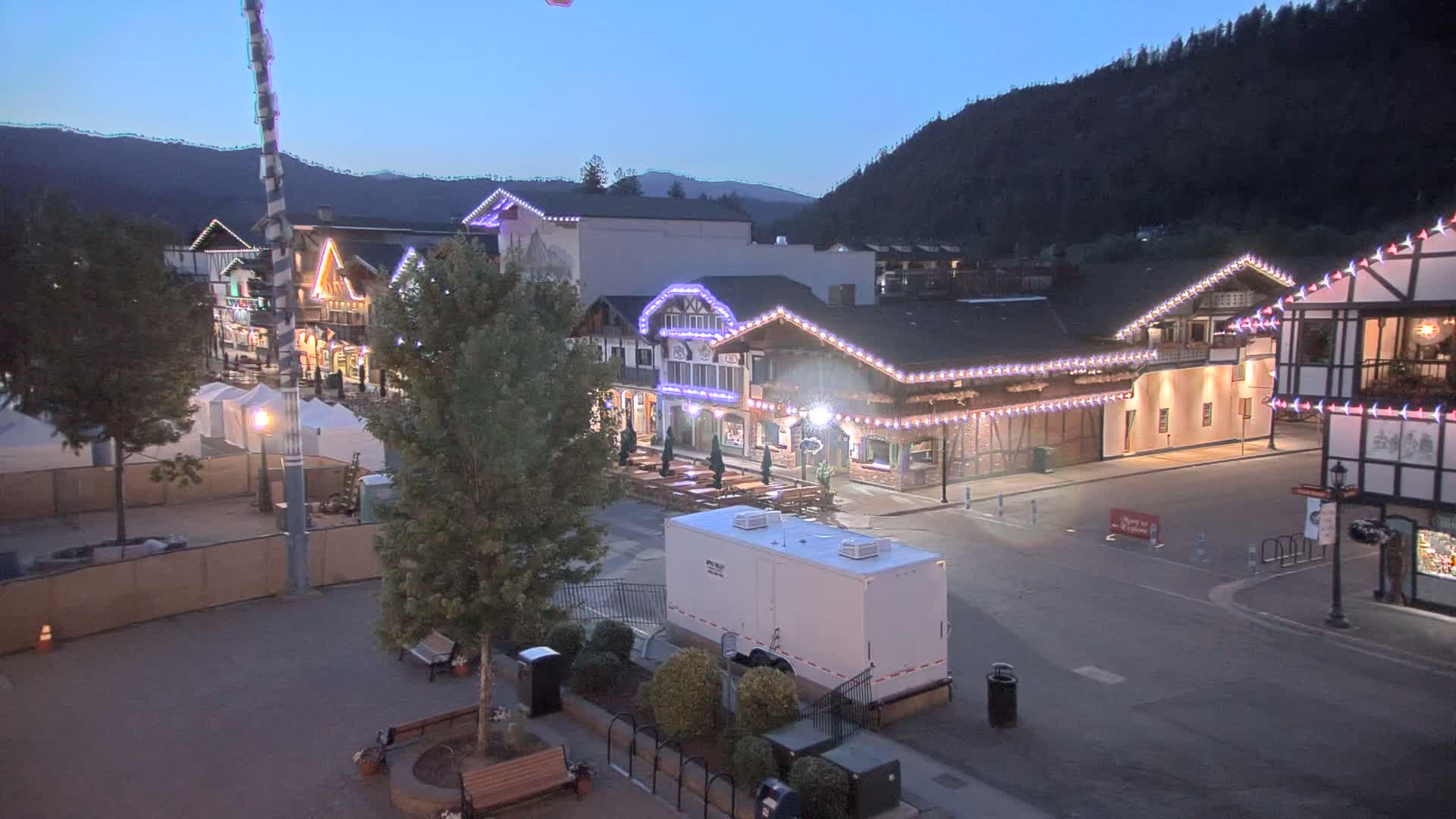 A town square at dusk, under a clear sky, features several buildings decorated with string lights, and a portable restroom sits in the center of the mostly empty square.