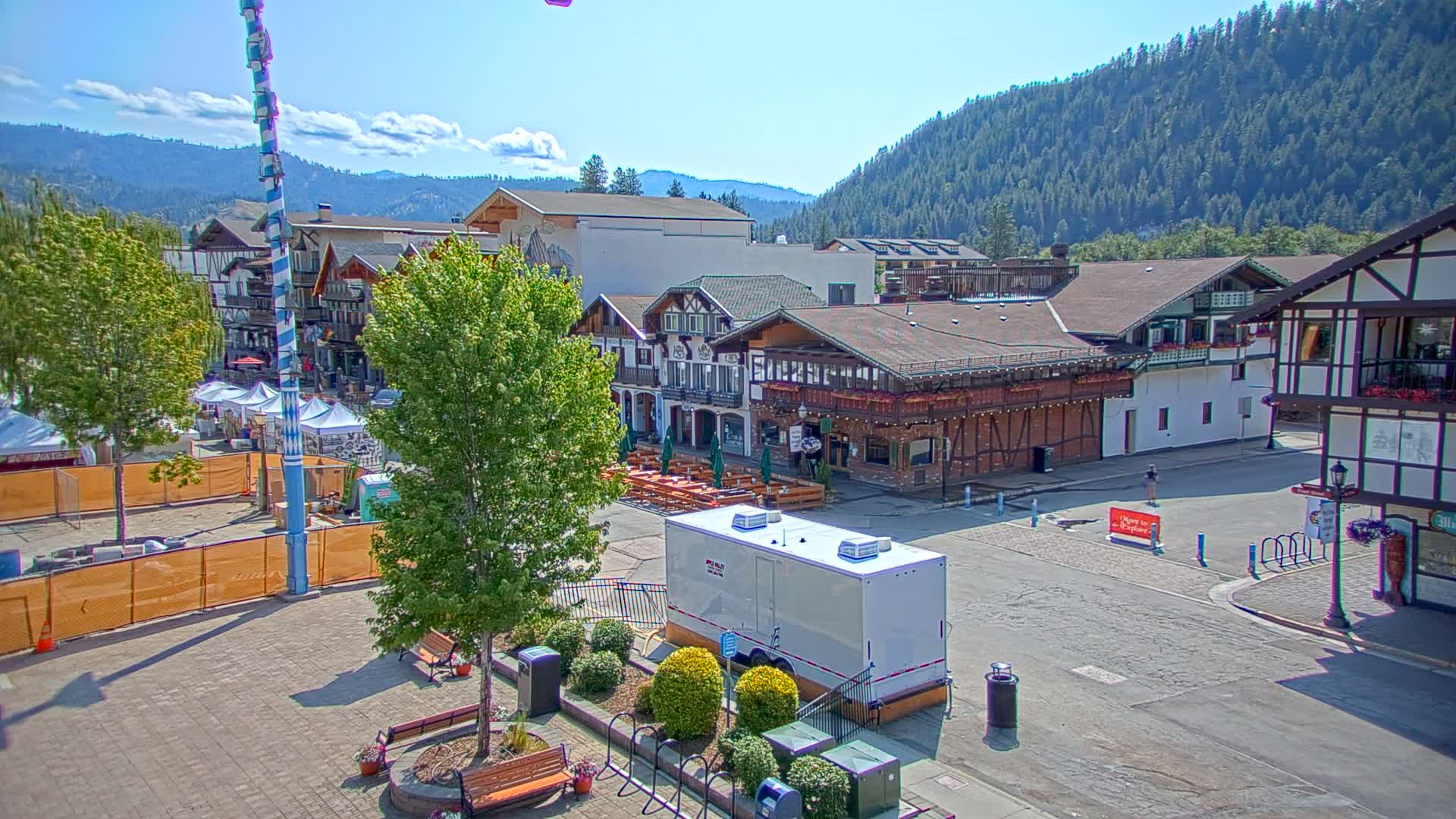 A town square with Bavarian-style buildings under a sunny sky, featuring a tall maypole and temporary structures suggestive of a festival or market.