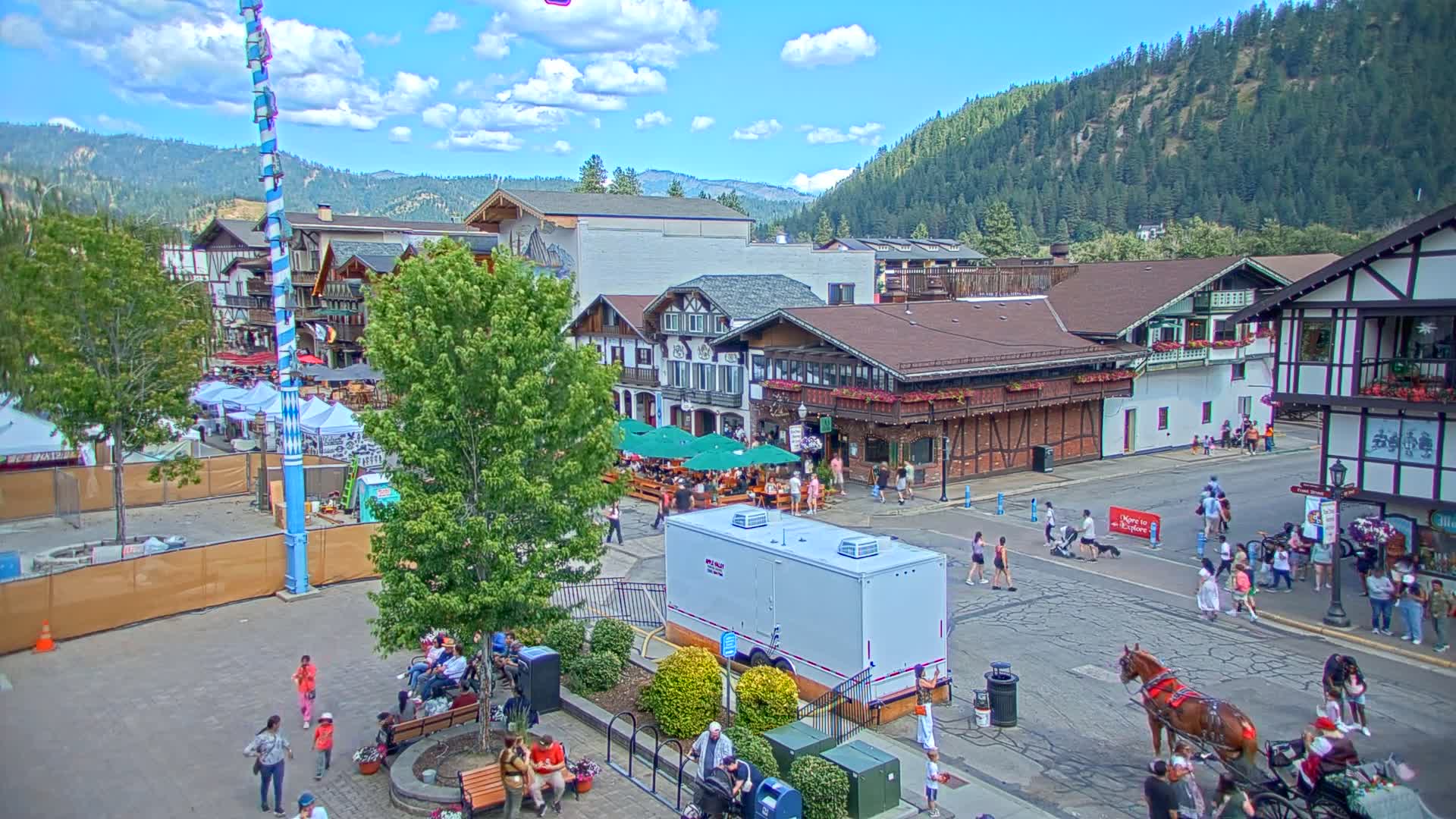 A town square with Bavarian-style buildings under a sunny sky, featuring a tall maypole and temporary structures suggestive of a festival or market.