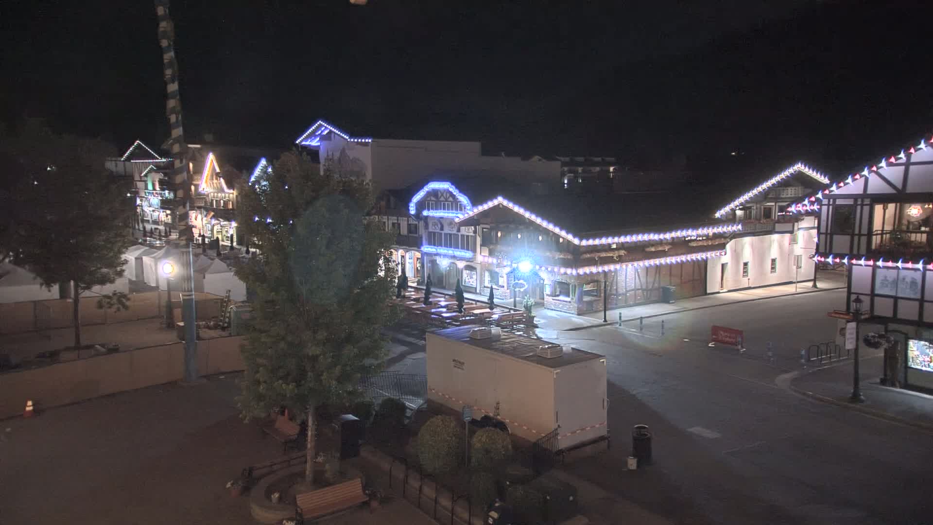 A nighttime, outdoor scene shows a plaza with several buildings decorated with string lights, some tents, trees, and a large trailer.