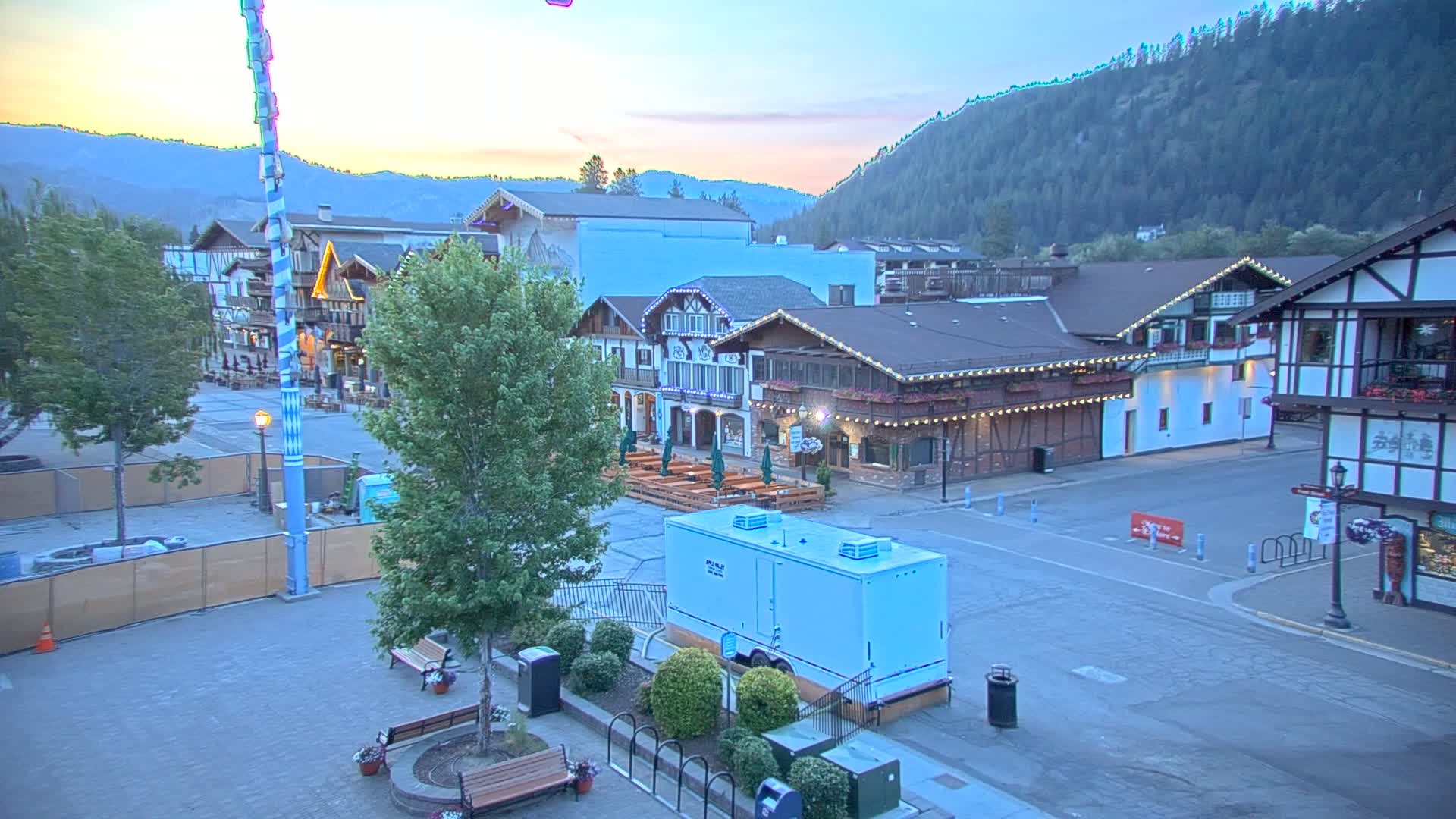 A mostly empty town square with Bavarian-style buildings at dusk, under a clear sky, surrounded by a mountain range.