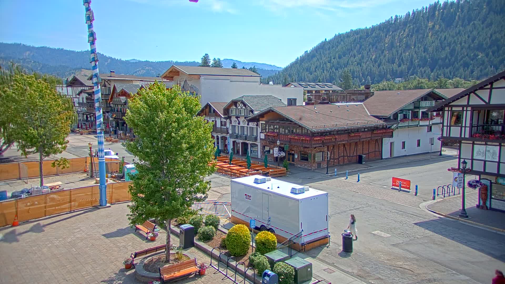 A paved town square with Bavarian-style buildings, trees, and temporary fencing under a sunny sky, backed by a mountain range.