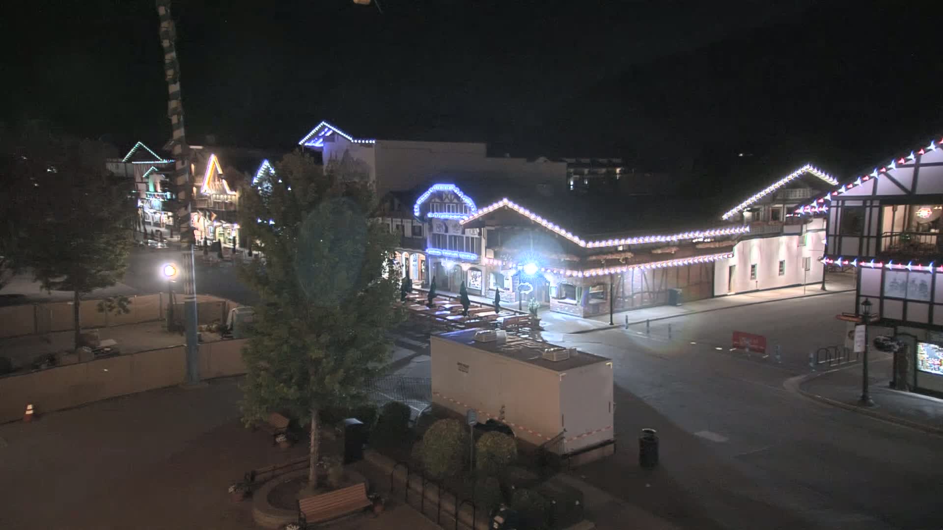 A nighttime, outdoor scene shows a row of illuminated buildings with festive lights, a mostly empty street, and a few trees.