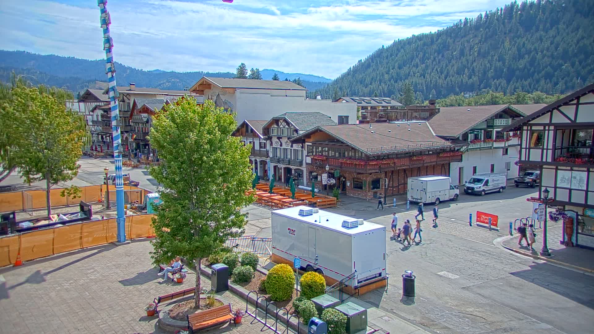 A town square with Bavarian-style buildings, a maypole, and several people walking under a partly cloudy sky.