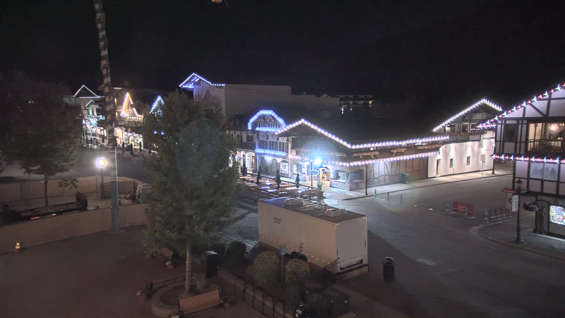 A nighttime, high-angle view shows a row of festively lit buildings with outdoor seating, a large trailer, and several trees in a mostly empty plaza.