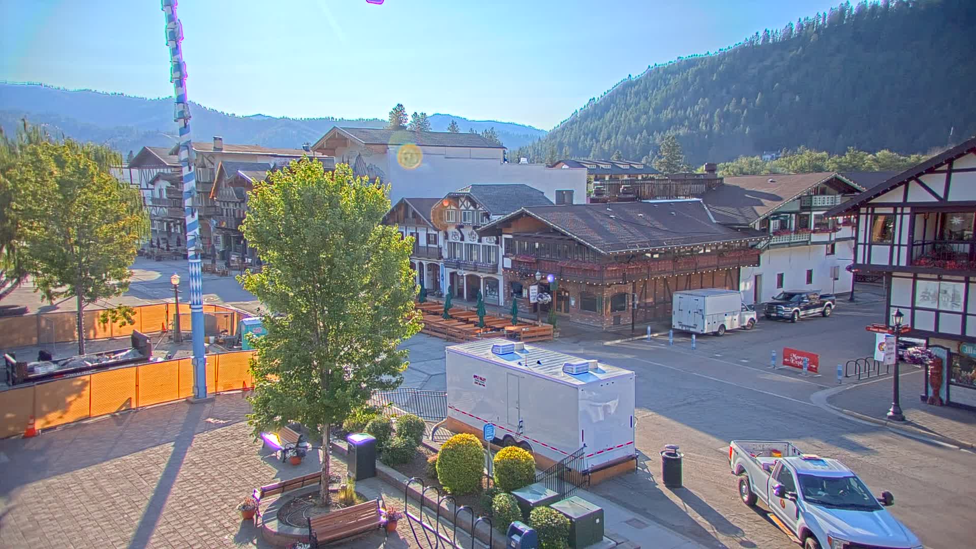 A Bavarian-style town square, under sunny skies, features several buildings, parked vehicles, and temporary restroom facilities, with a large, striped pole in the center and mountains in the background.