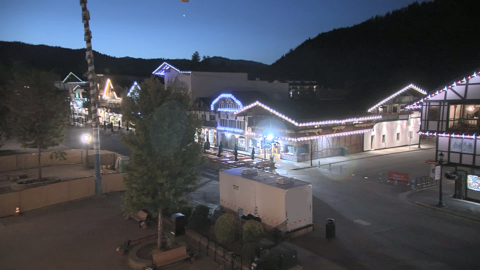 A nighttime view of a town square with several buildings decorated with string lights, under a dark blue, clear sky.